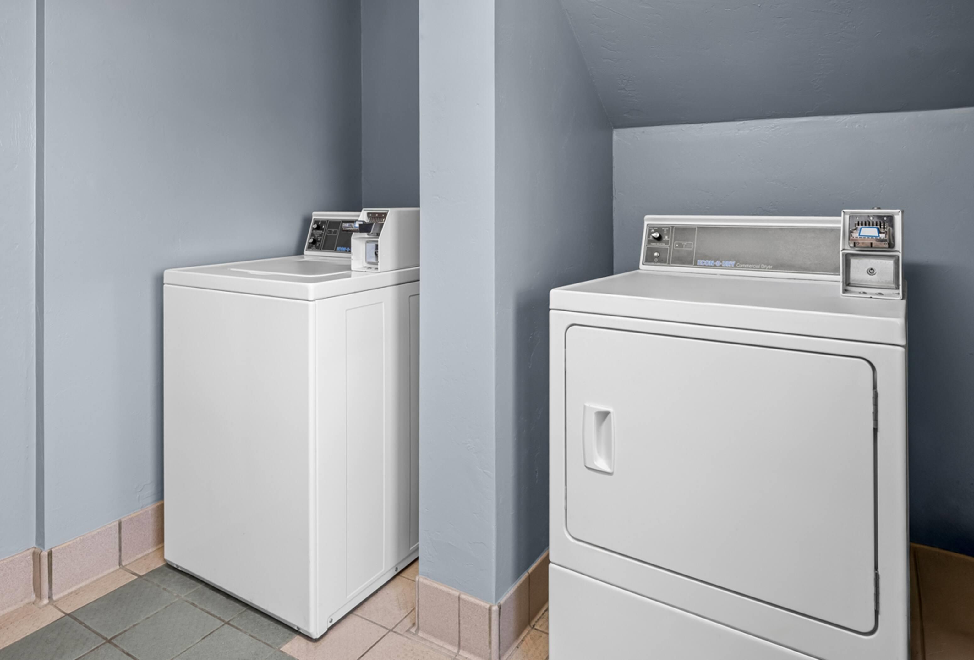 Une buanderie équipée de deux machines à laver et deux sèche-linge superposés, avec un sol carrelé et des murs de couleur bleu clair.