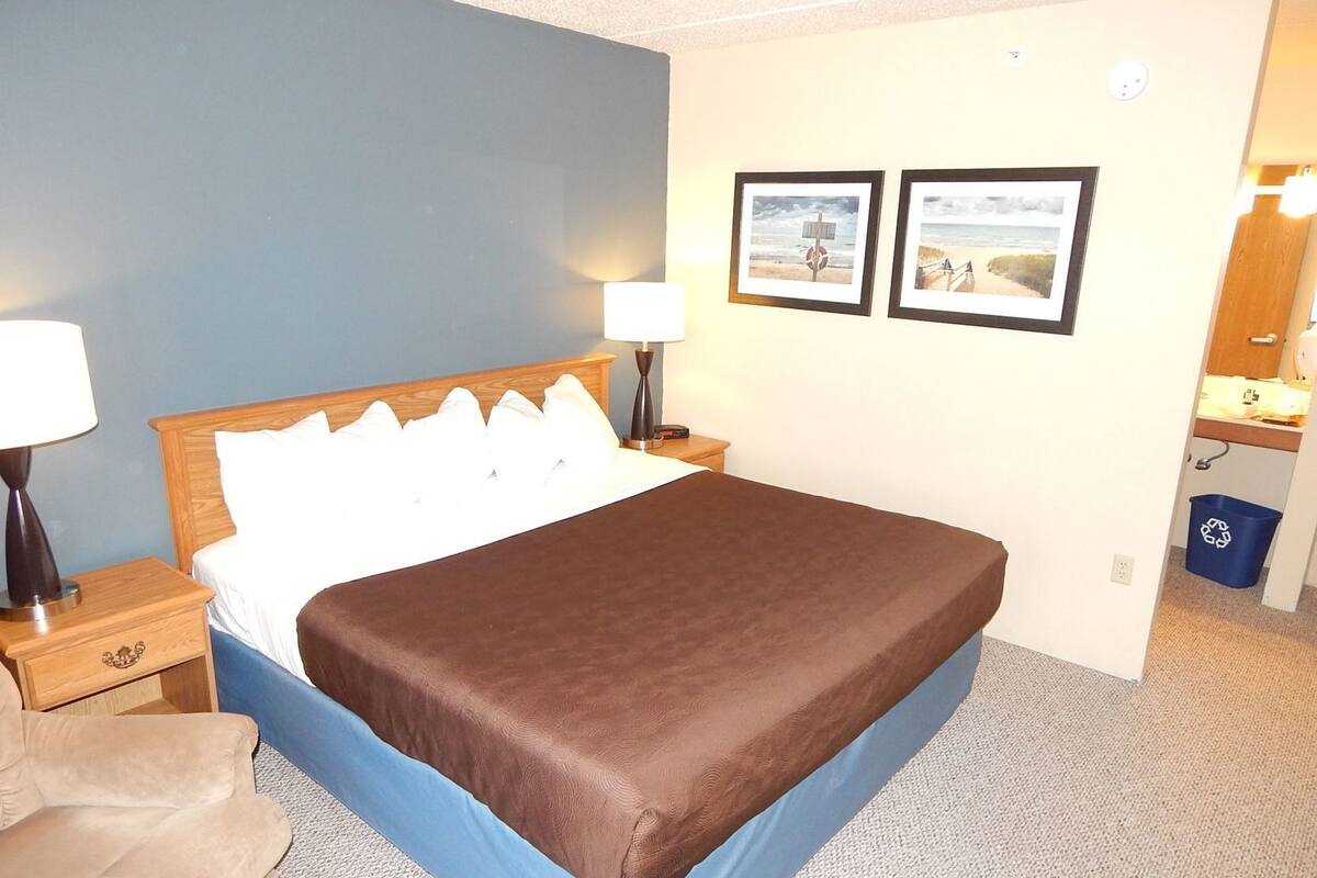 Een hotelkamer met een bed, twee bedlampen, een nachtkastje, een stoel, een blauwe prullenbak en twee foto's in lijsten aan de muur.