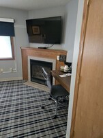 A room with a fireplace, a television mounted above it, a desk with a chair, and a patterned carpet.