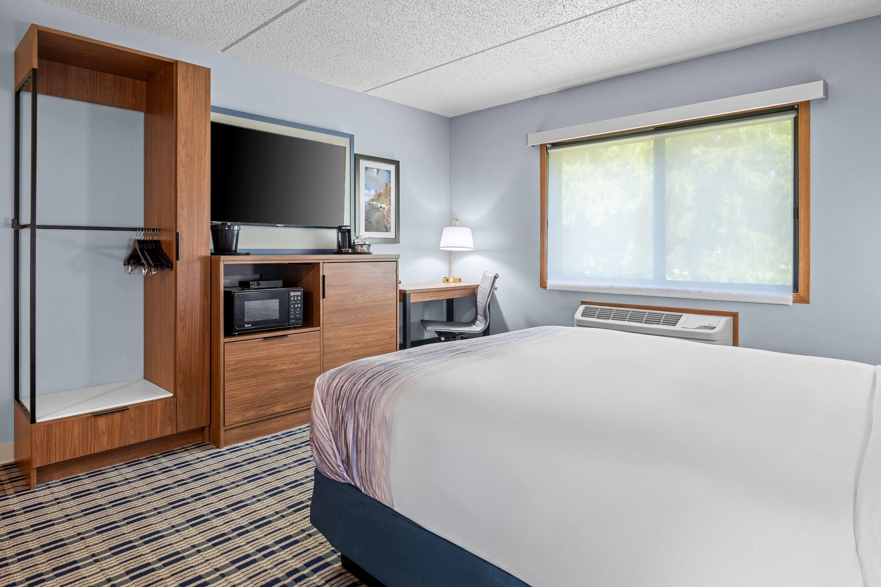 A hotel room with a bed, a desk with a lamp, a television, and a window with blinds.