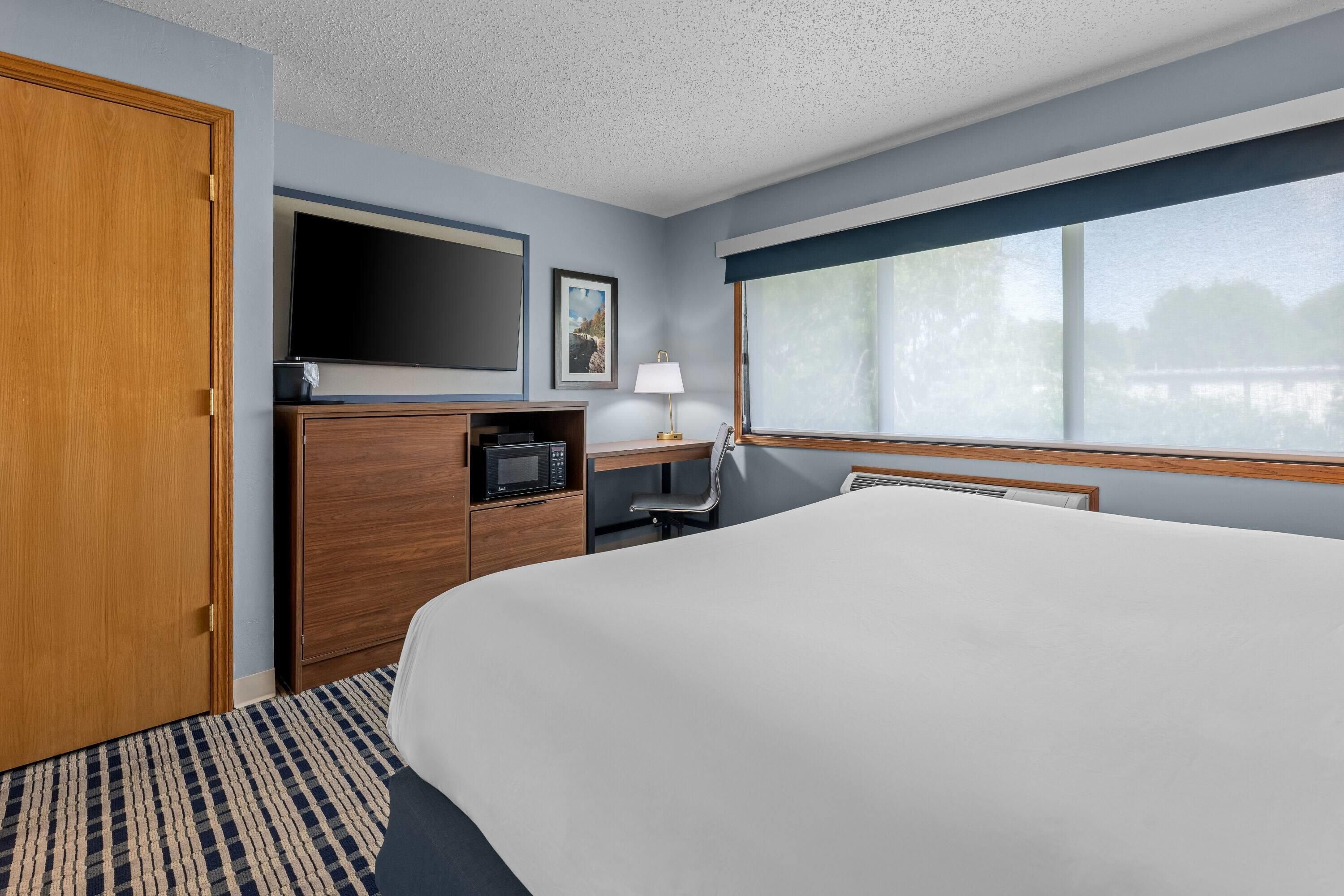 A hotel room with a large bed, a flat-screen TV, a microwave, a desk with a lamp, and a window with a view.