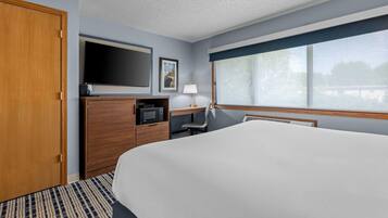 A hotel room with a large bed, a flat-screen TV, a microwave, a desk with a lamp, and a window with a view.