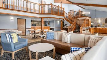 A spacious hotel lobby with a high ceiling, a central seating area with sofas and chairs, a staircase connecting two floors, and a reception desk.