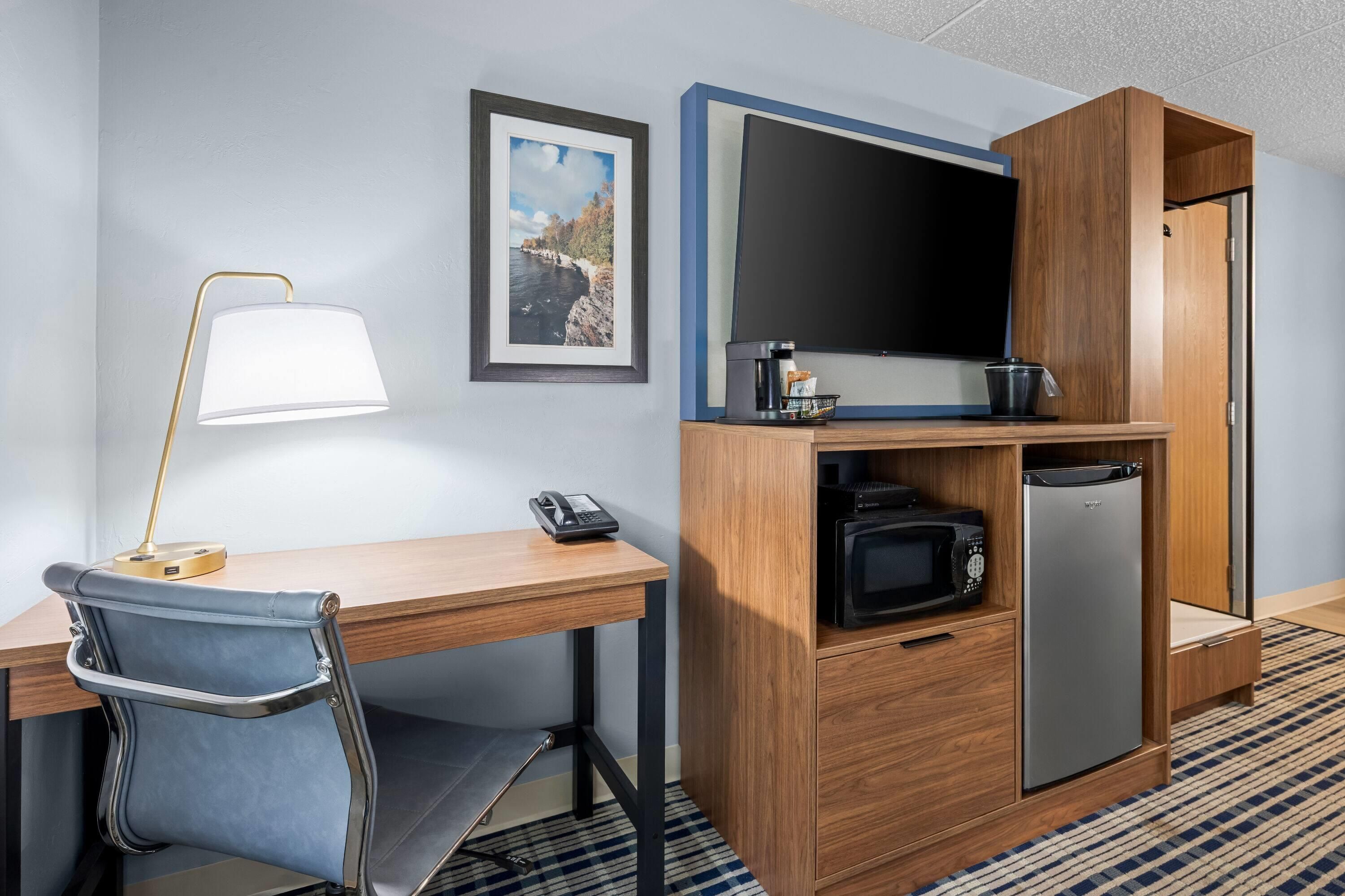 A hotel room with a desk, chair, lamp, TV, coffee maker, phone, microwave, and refrigerator.