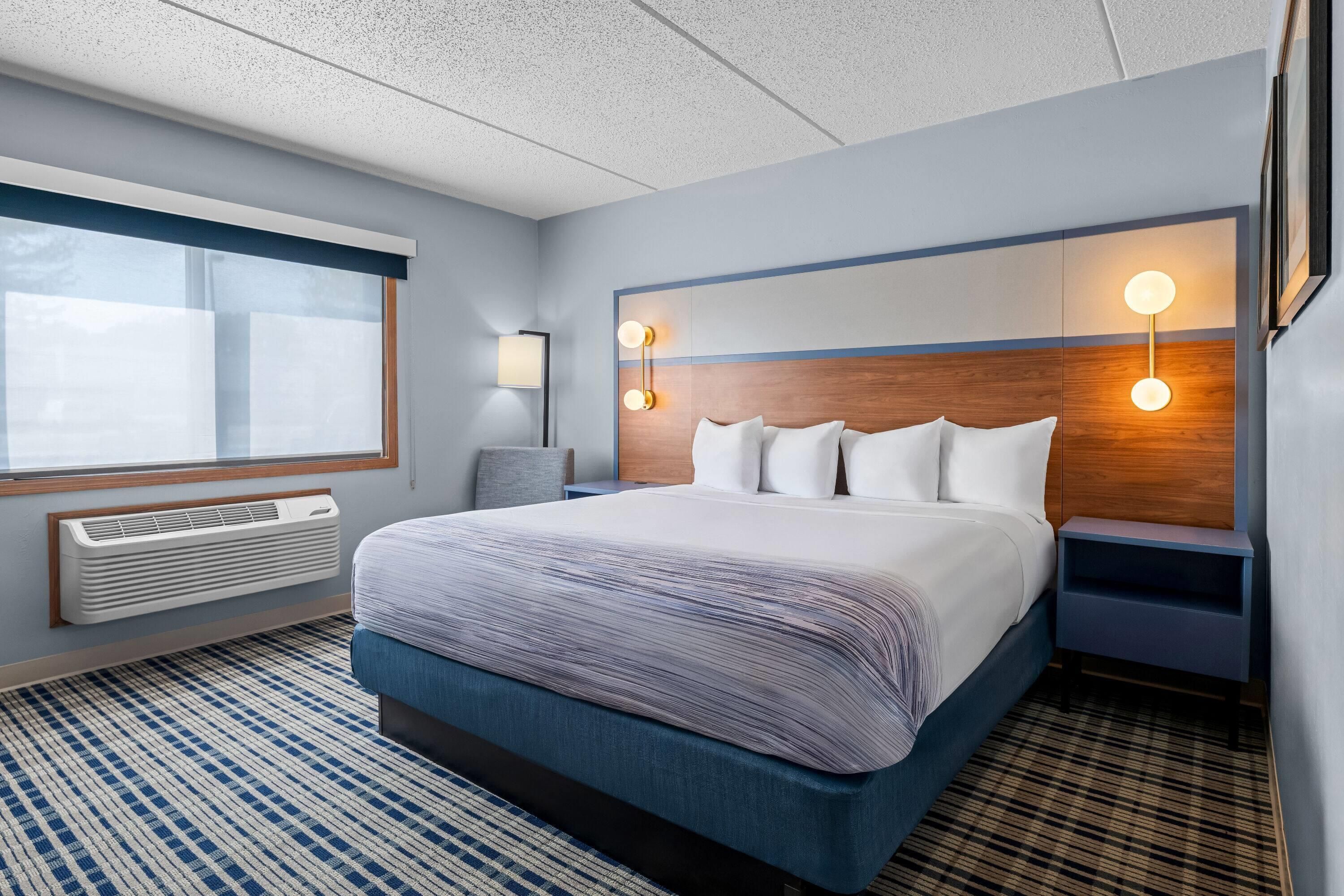 A hotel room with a large bed, a window, a wall-mounted lamp, and a bedside table.