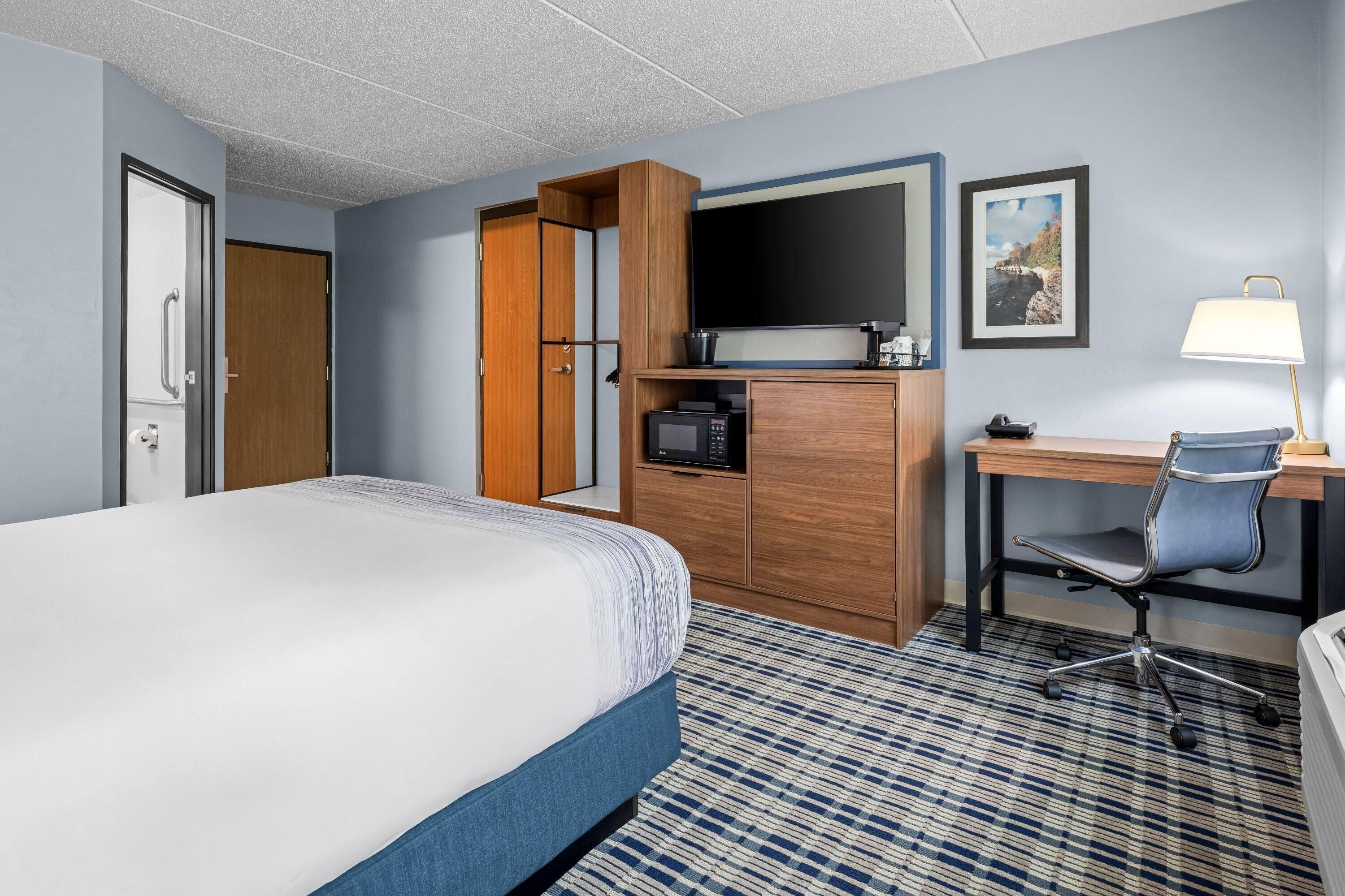 Quarto de hotel com cama, escrivaninha, cadeira, televisão e cafeteira.