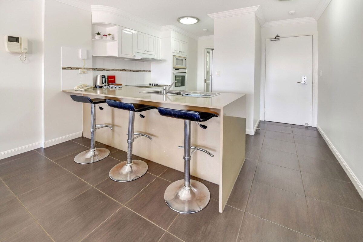 A modern kitchen with a central island, three bar stools, and a built-in oven.