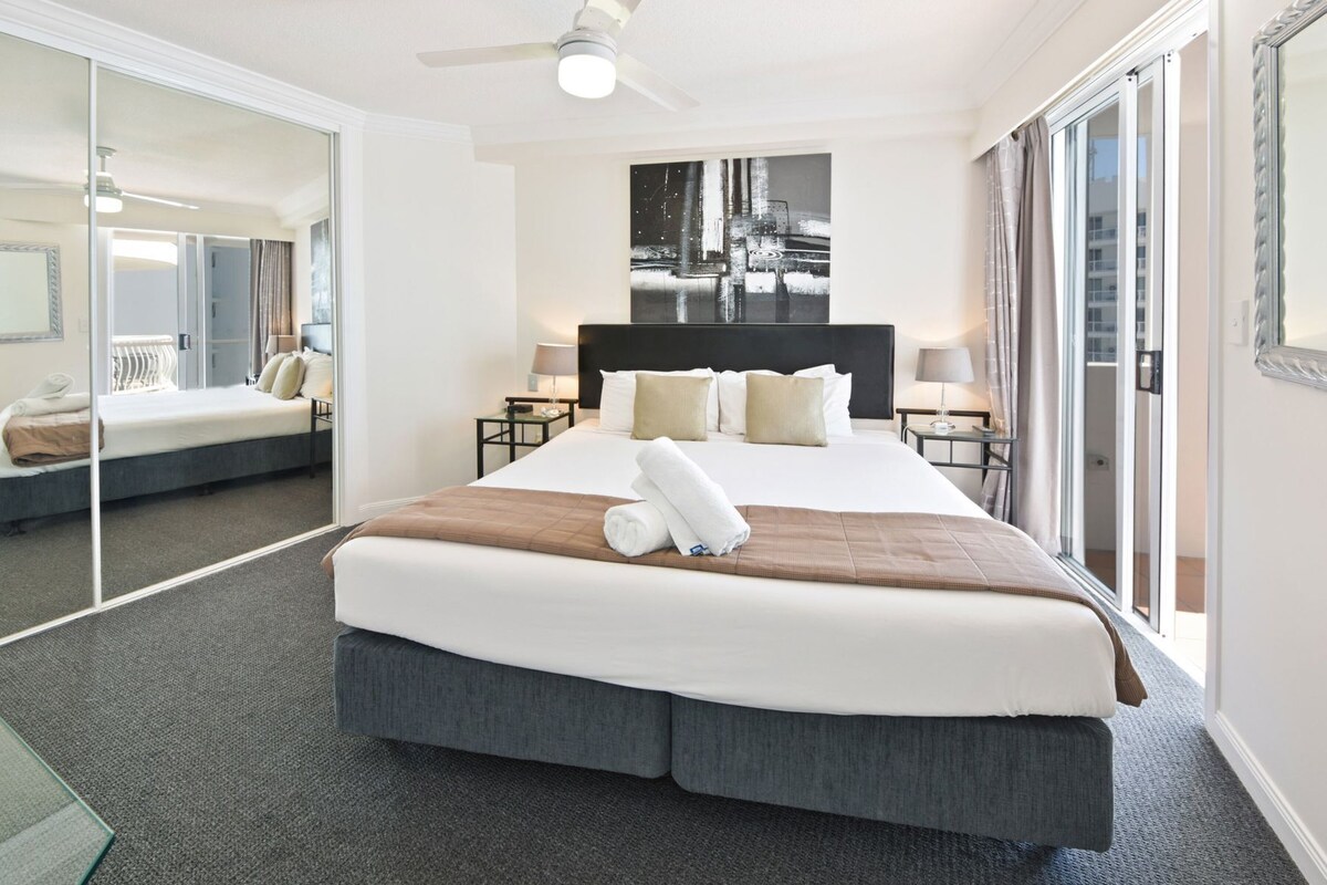 A hotel room with a large bed, a ceiling fan, a cityscape painting, and a sliding glass door.
