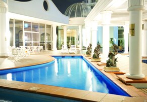 Una piscina con columnas blancas y sillas, rodeada por una zona cubierta con puertas de cristal.