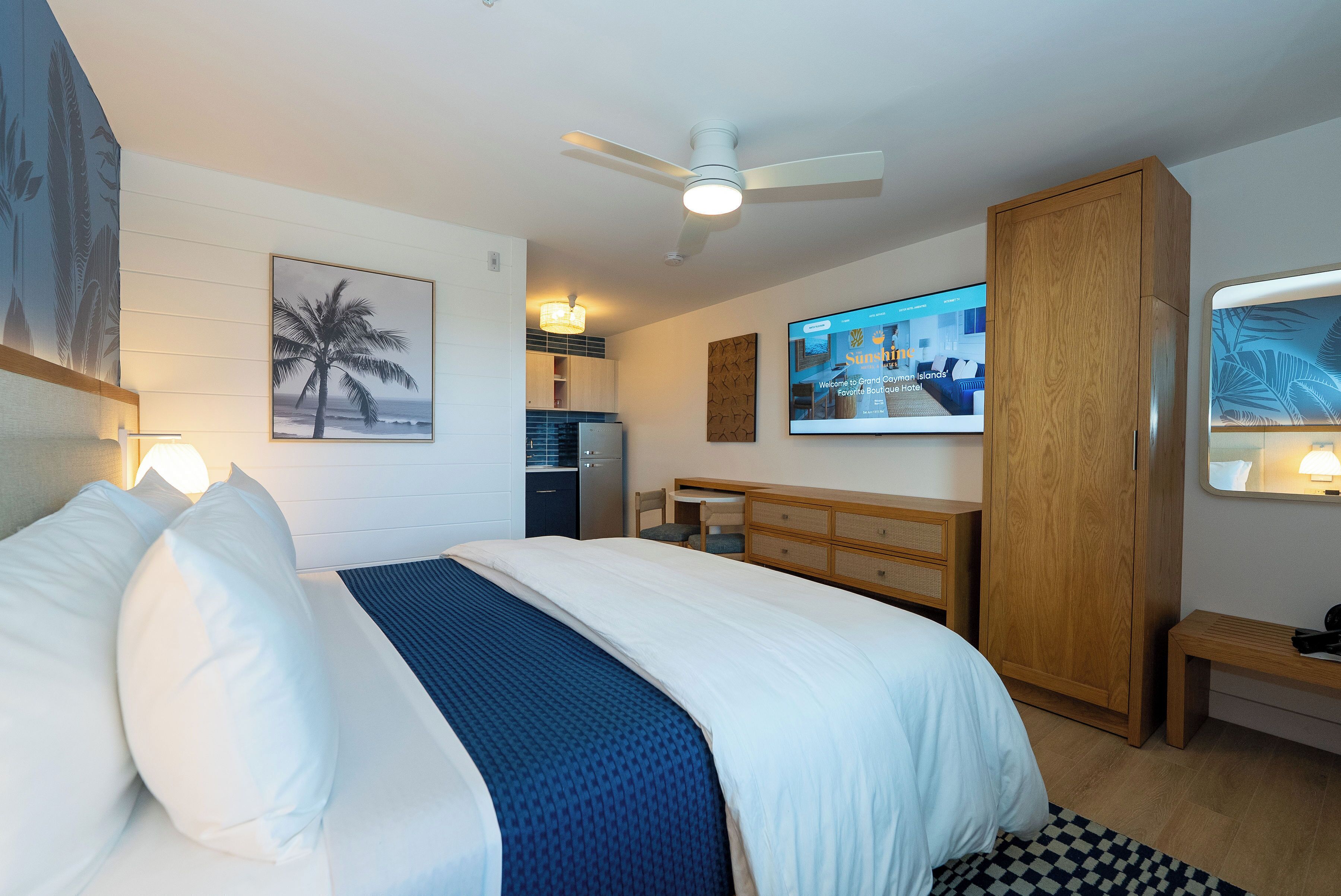 A hotel room with a bed, a television, a ceiling fan, a minibar, and a wall-mounted TV displaying a hotel website.
