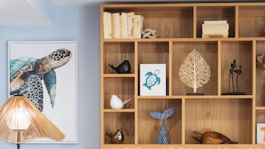 A modern living room with a grey sofa, two round blue ottomans, a wooden bookshelf with various decorative items, and a framed sea turtle artwork.