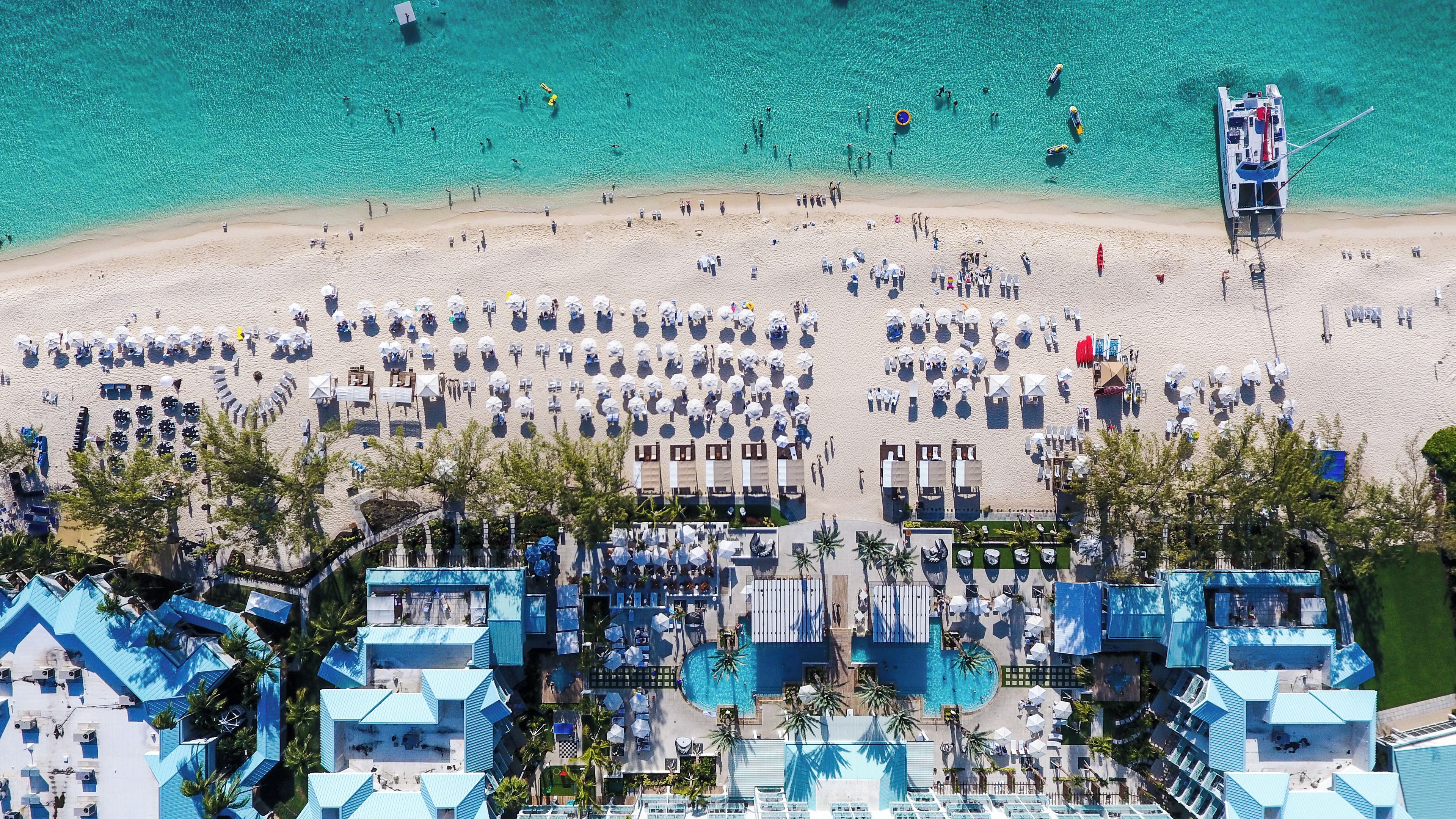 Vista aérea de un complejo turístico en la playa con varias piscinas, tumbonas y un barco grande amarrado a la orilla.