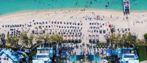 Vista aérea de un complejo turístico en la playa con varias piscinas, tumbonas y un barco grande amarrado a la orilla.