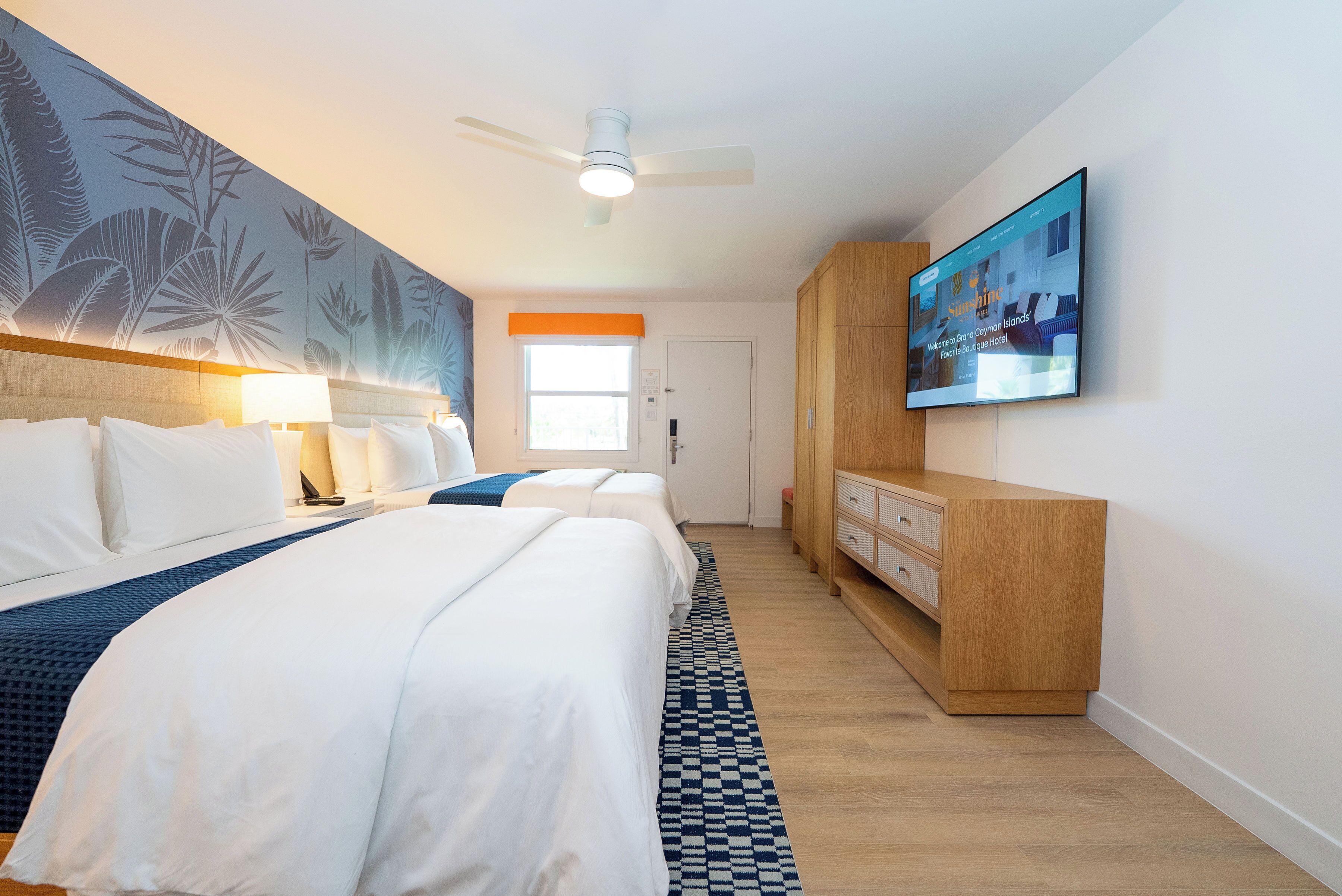 A hotel room with two beds, a ceiling fan, a large window, and a television on the wall.