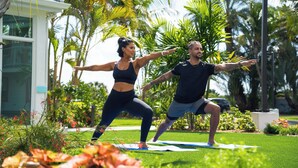 Two people exercising on a mat in a garden with palm trees and a building in the background. - The Sunshine Hotel and Suites (Seven Mile Beach)