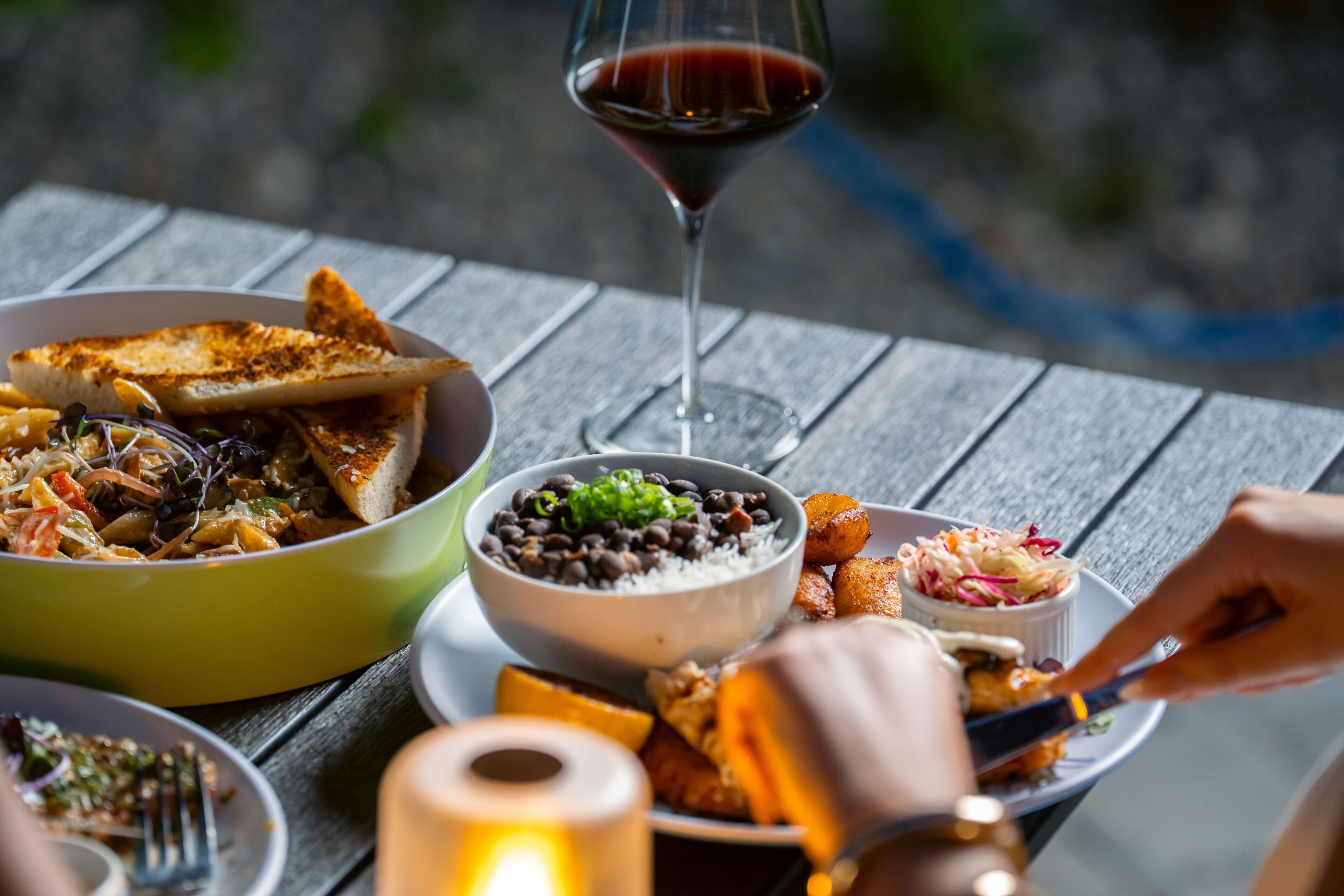 Una mesa de comedor con diversos platos, una copa de vino tinto y la mano de una persona sosteniendo un tenedor.