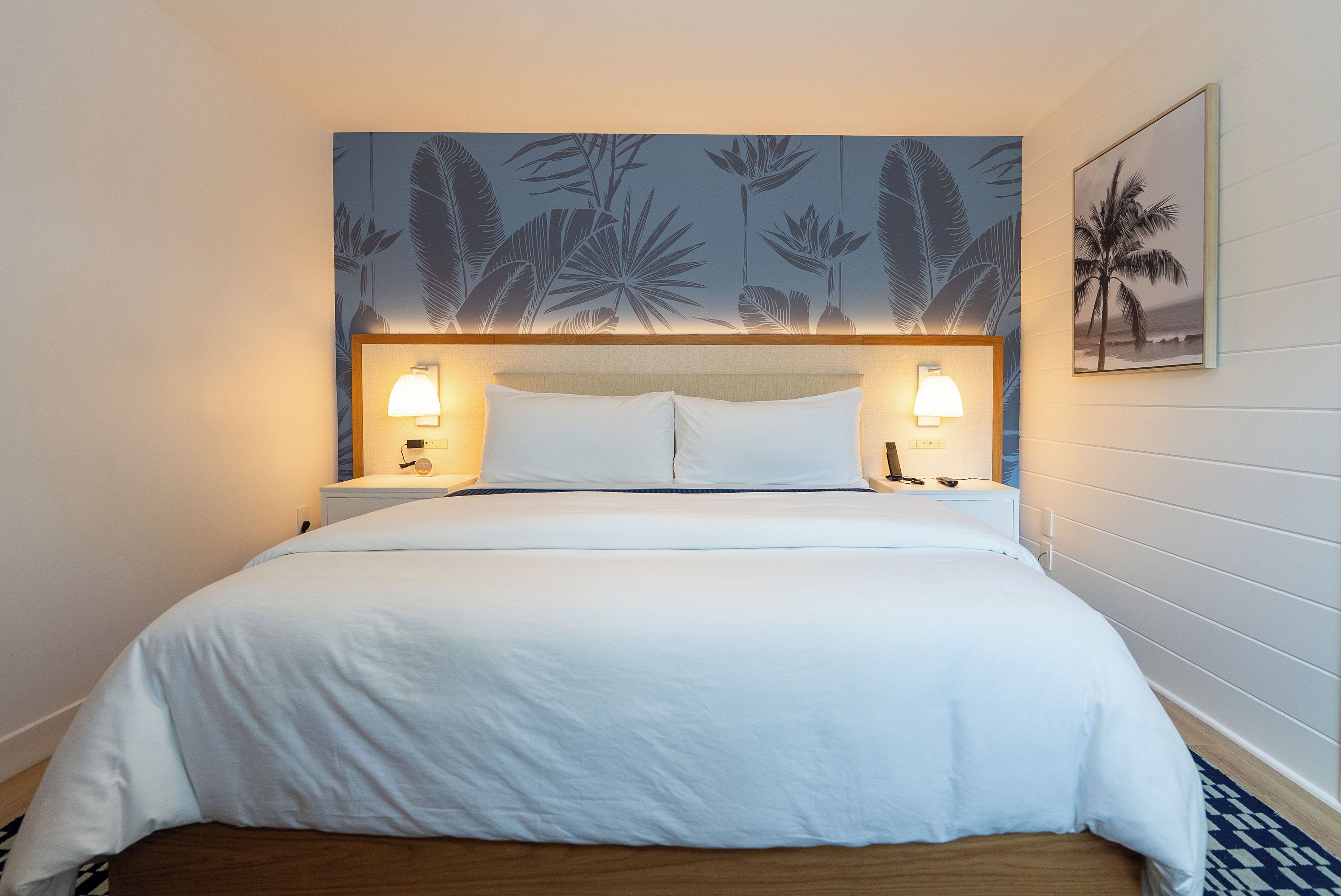 Habitación de hotel con cama, dos lámparas de noche, cabecera con diseño de hojas y un cuadro en la pared.