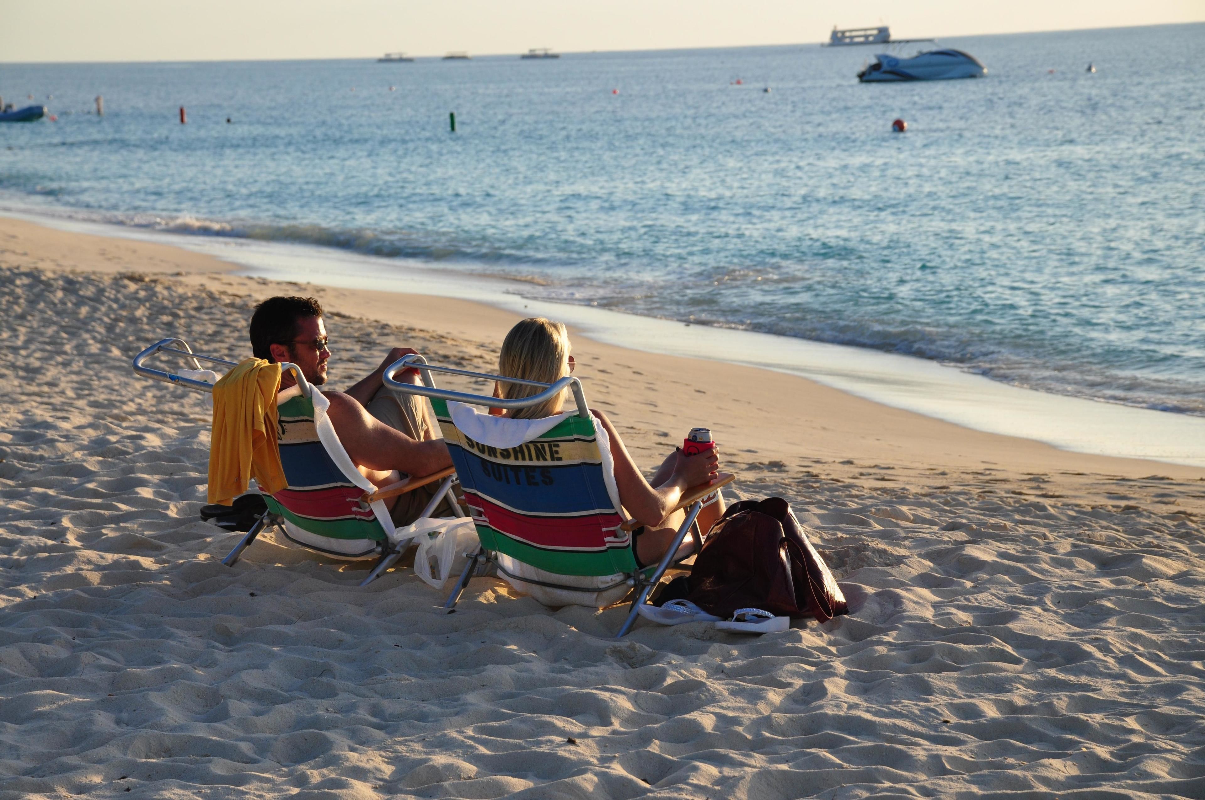 Two people sitting on beach chairs on a sandy beach with a view of the ocean and boats in the distance.