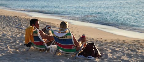 Two people sitting on beach chairs on a sandy beach with a view of the ocean and boats in the distance.