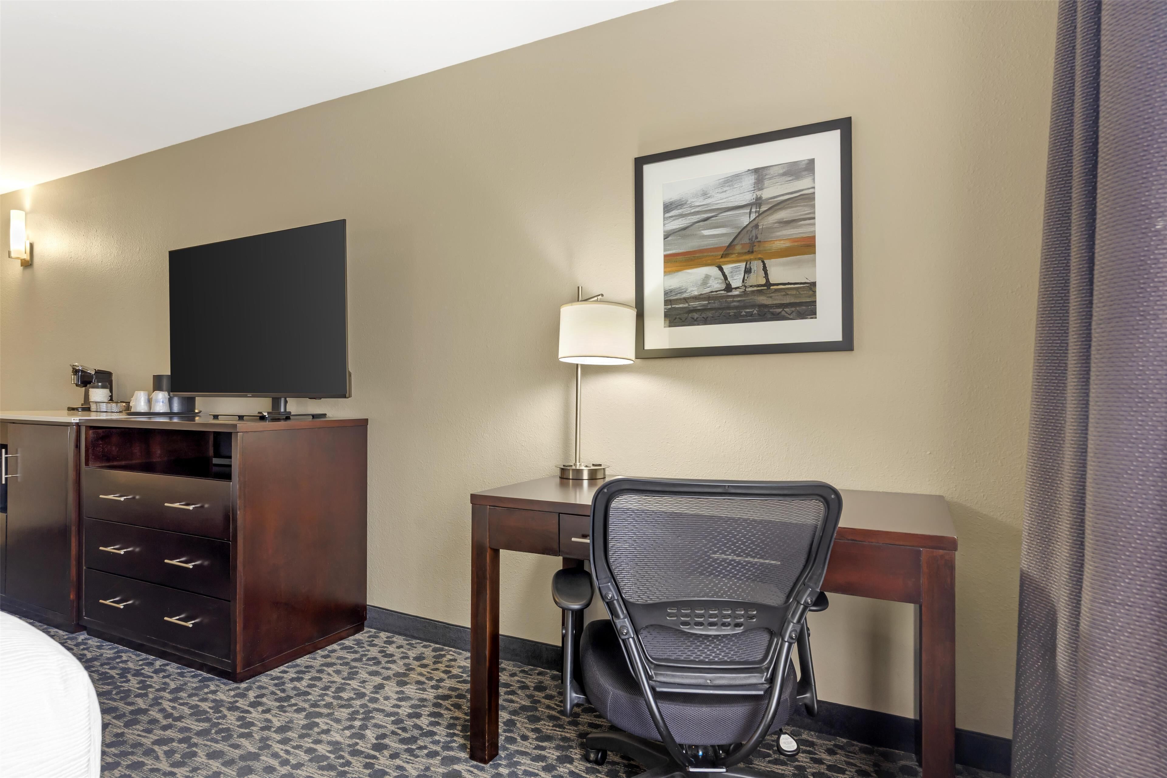 A hotel room with a bed, a desk with a chair, a television, a lamp, and a framed picture on the wall.