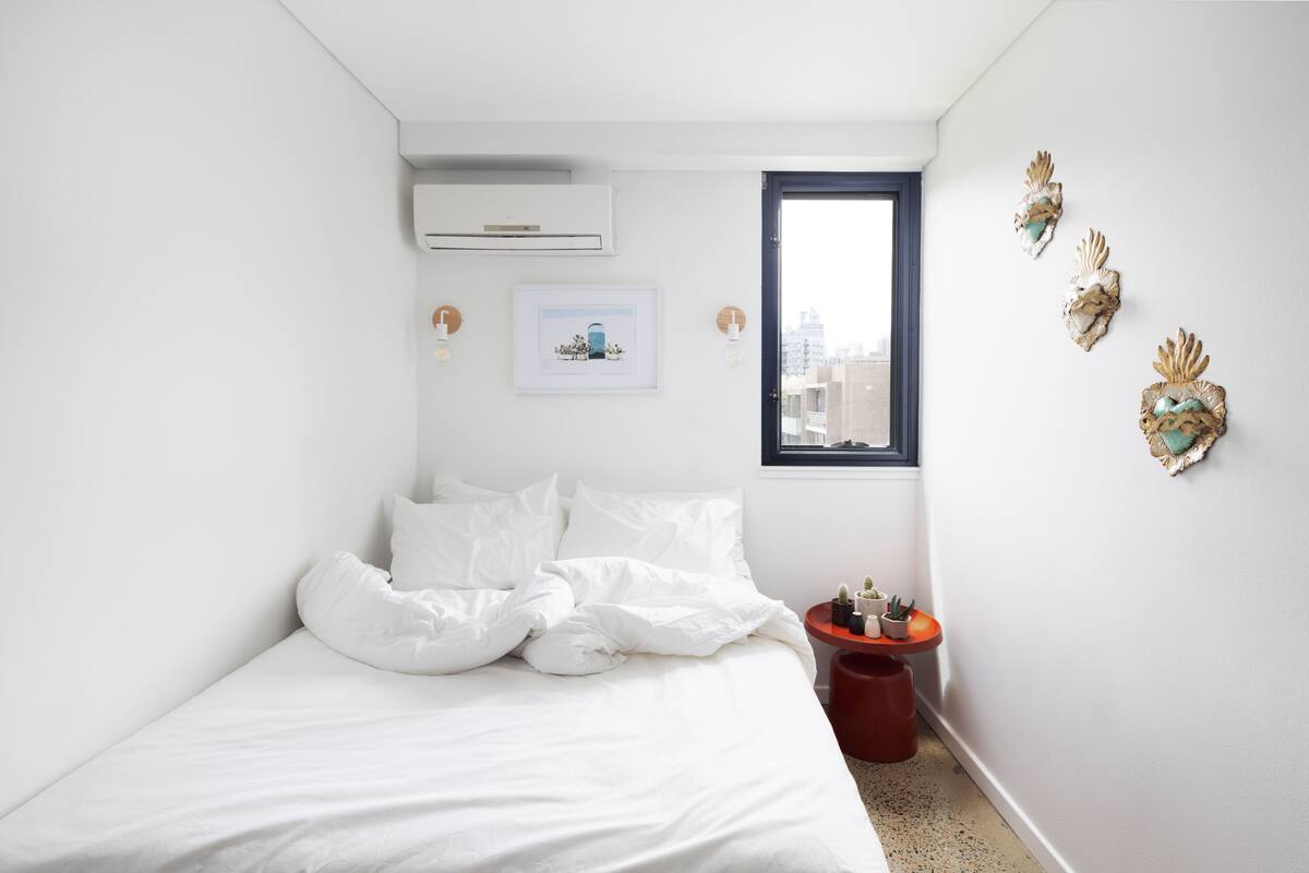 A bedroom with a bed, a window, a wall-mounted air conditioner, a framed picture, and decorative wall art.