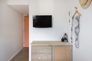 Una stanza con una TV a schermo piatto montata al muro, un comò in legno con cassetti, un mobiletto in vimini e un cappello appeso a un appendiabiti in legno.