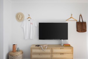 Huoneessa on seinälle asennettu televisio, puinen lipasto laatikoineen, punottu jakkara sekä hattu ja laukku, jotka roikkuvat seinällä.