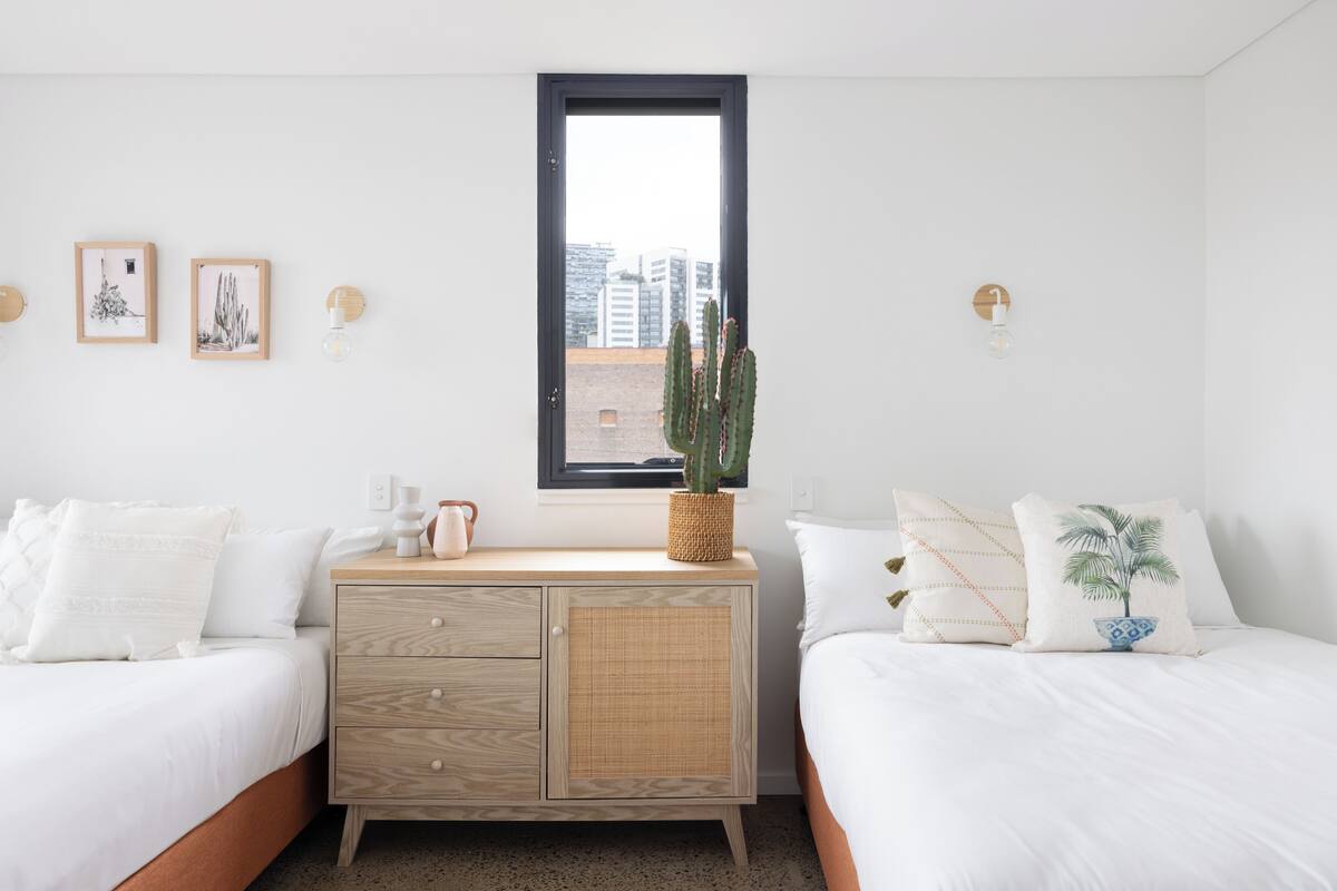 A bedroom with two beds, a wooden nightstand, a window, and wall-mounted light fixtures.