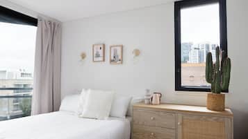 A bedroom with a bed, a dresser, a window with a view of buildings, and a cactus on the dresser.