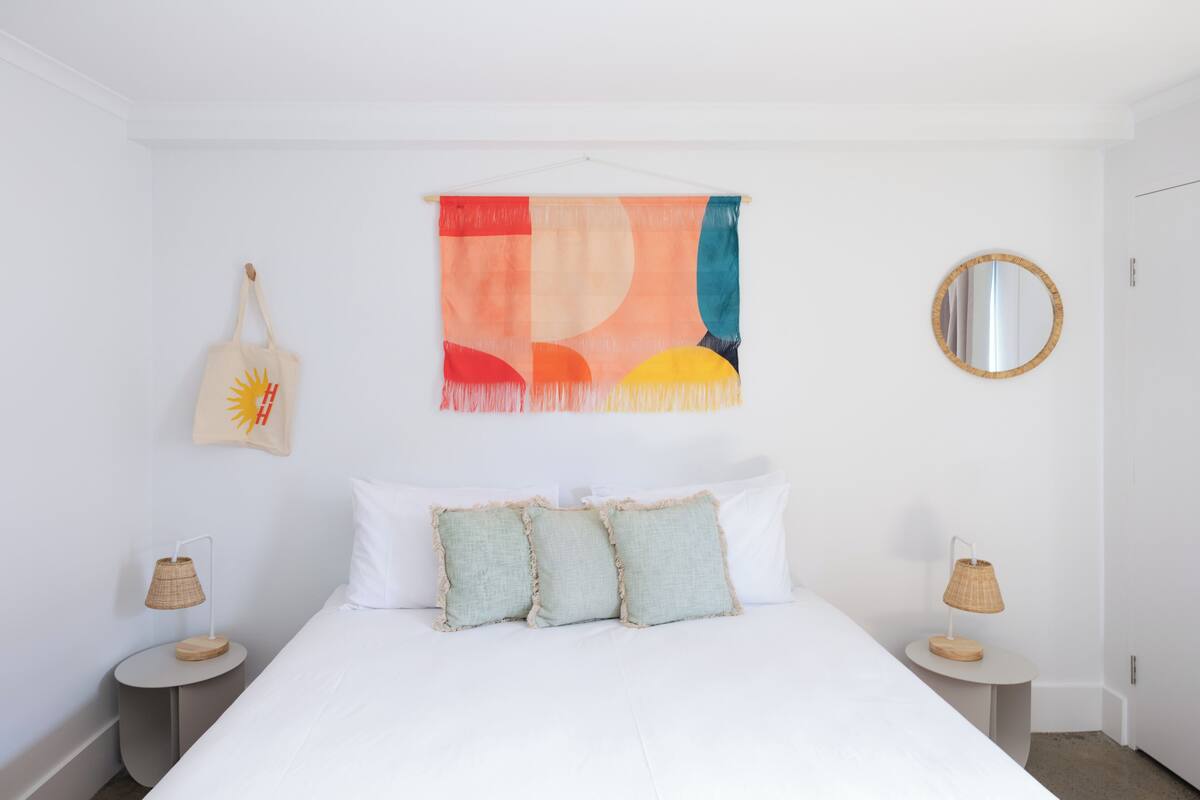 A bedroom with a bed, two bedside tables, a wall hanging, a hanging bag, and a round mirror.