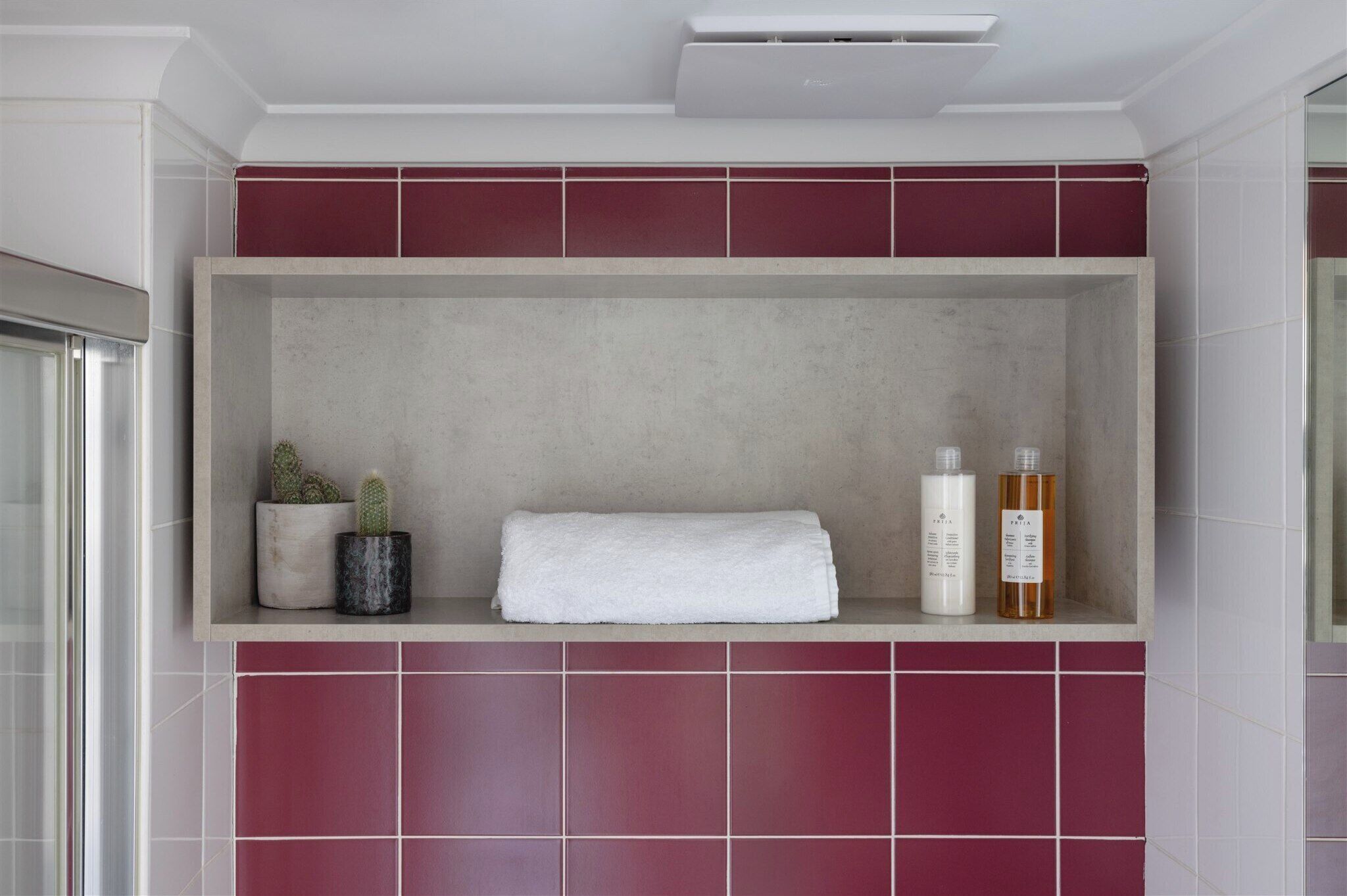 A bathroom with a built-in shelf holding a white towel, two bottles, and two potted plants.