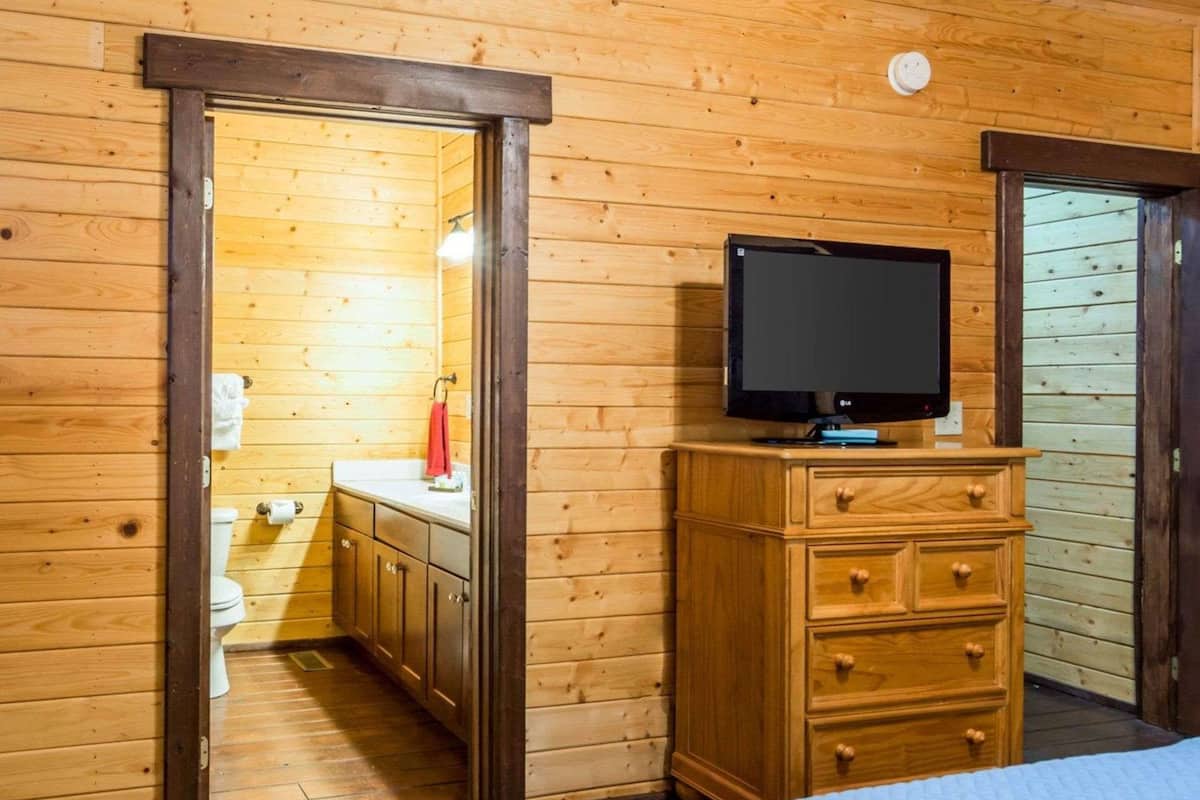 Une pièce aux murs lambrissés, avec un lit, un téléviseur posé sur une commode en bois, et une salle de bain visible à travers une porte ouverte.