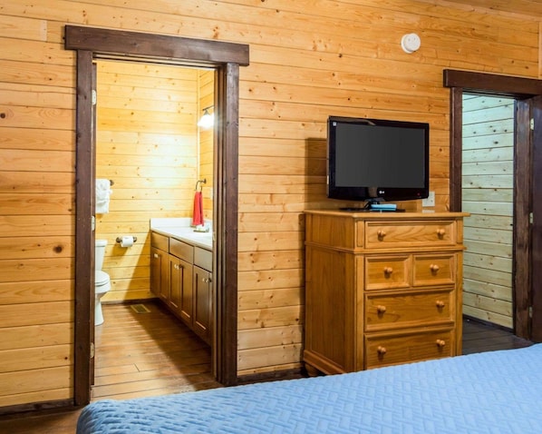 Una habitación con paneles de madera, una cama, un televisor sobre un aparador de madera y un baño visible a través de una puerta abierta.
