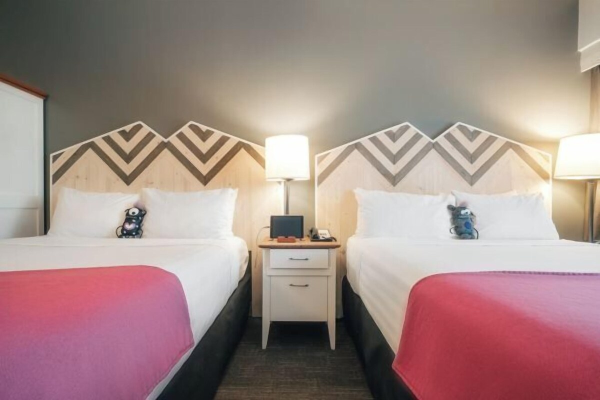 A hotel room with two beds, a nightstand with a lamp, and a small table with a phone and a book.