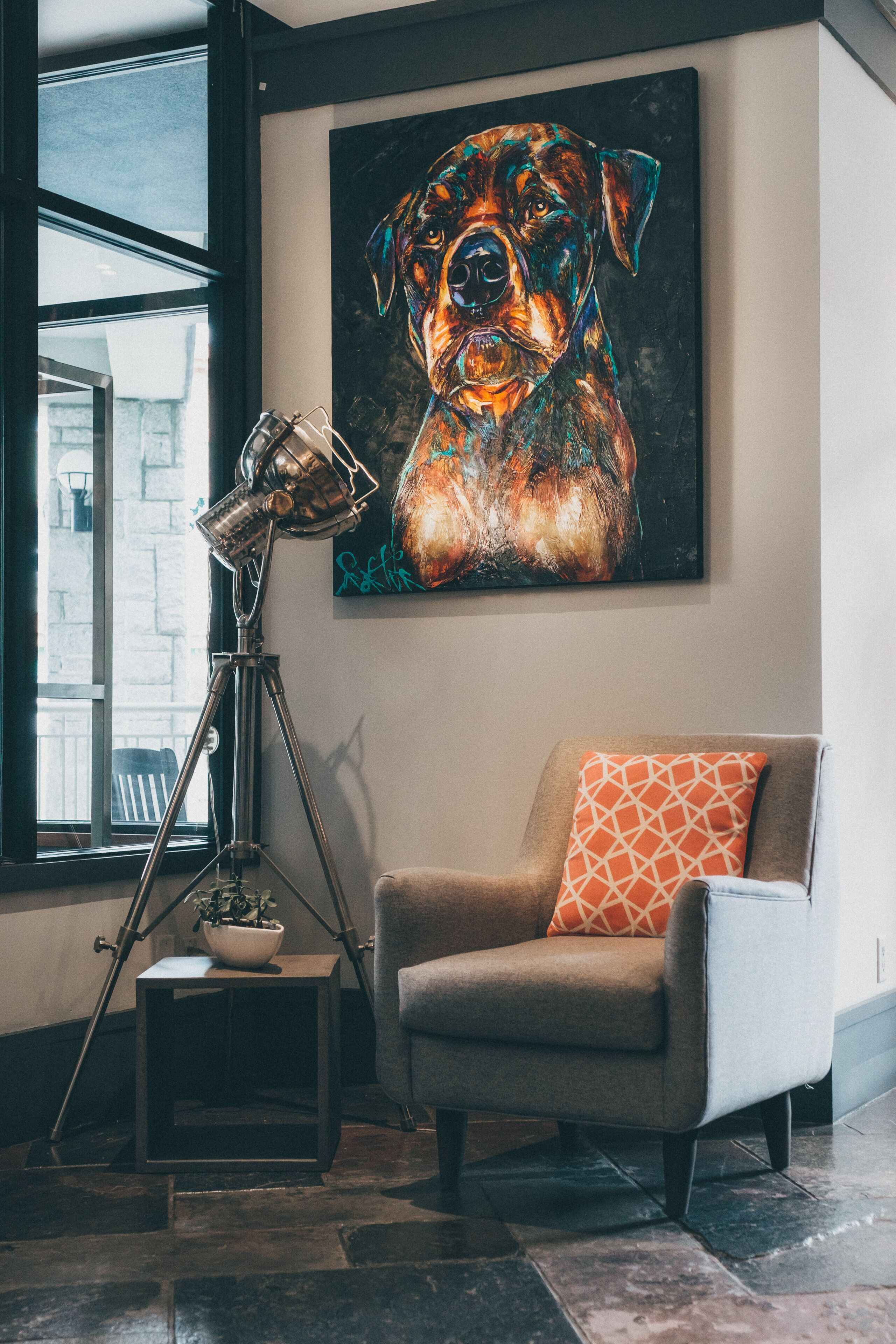 Een kamer met een groot raam, een statief met lamp, een stoel met oranje kussen en een schilderij van een hond aan de muur.