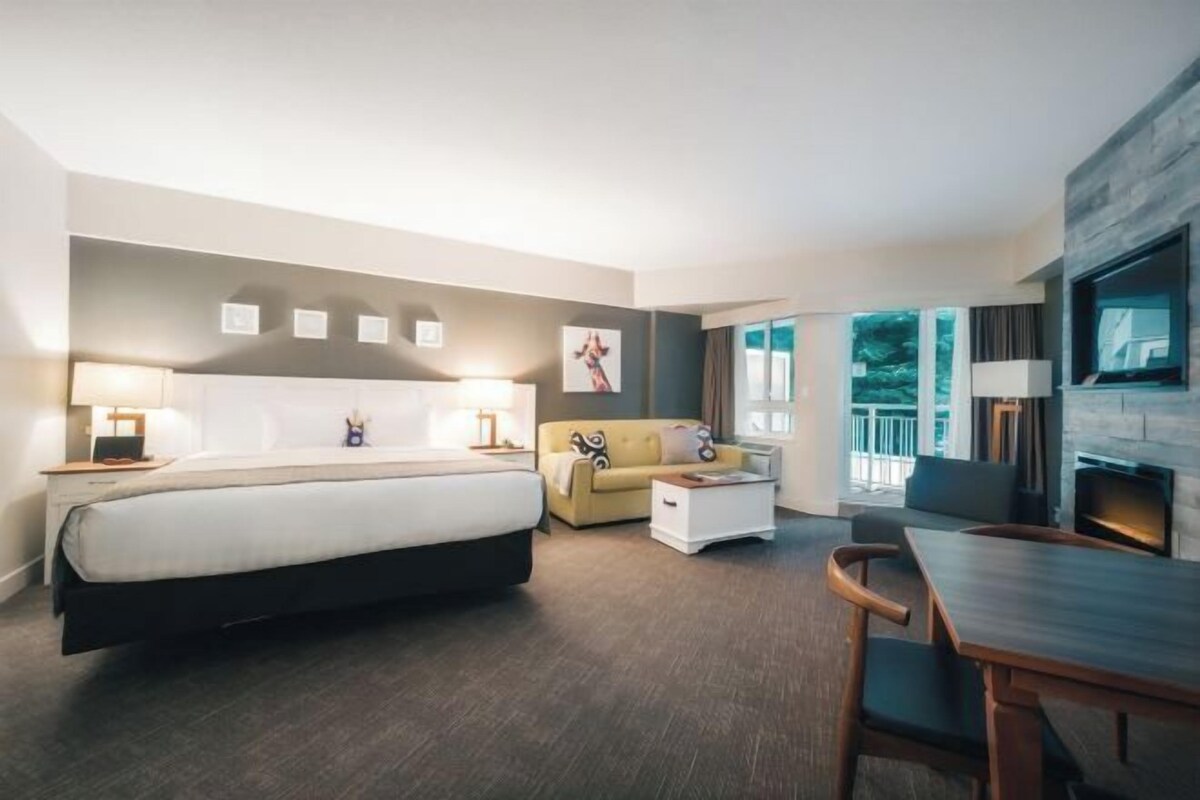 A modern hotel room with a bed, sofa, dining table, and a TV.