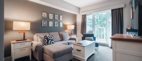 A modern hotel room with a grey sofa, a small white table, a TV, and a balcony with curtains.