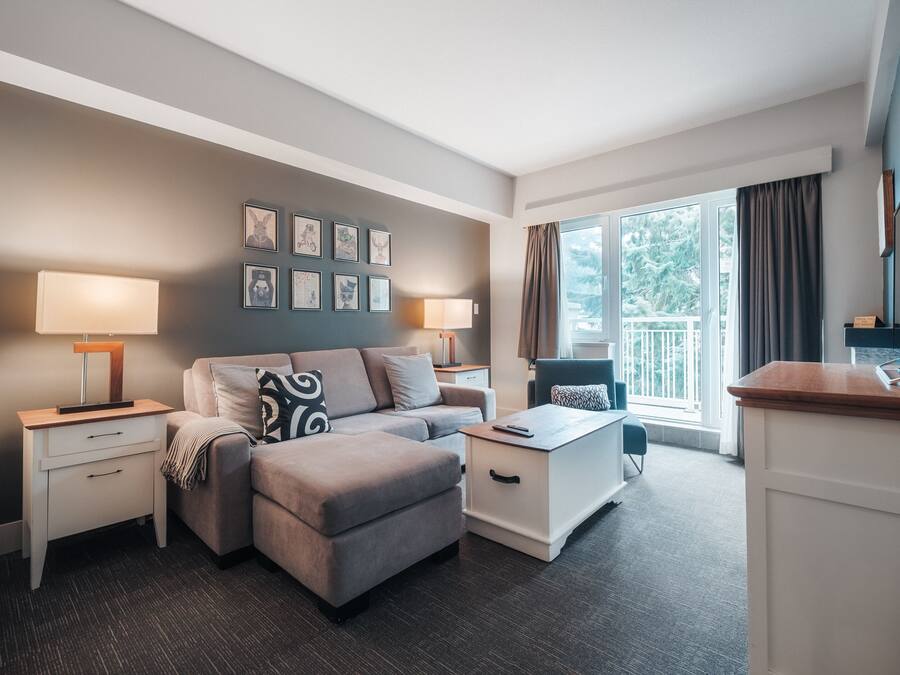 A modern hotel room with a grey sofa, a small white table, a TV, and a balcony with curtains.