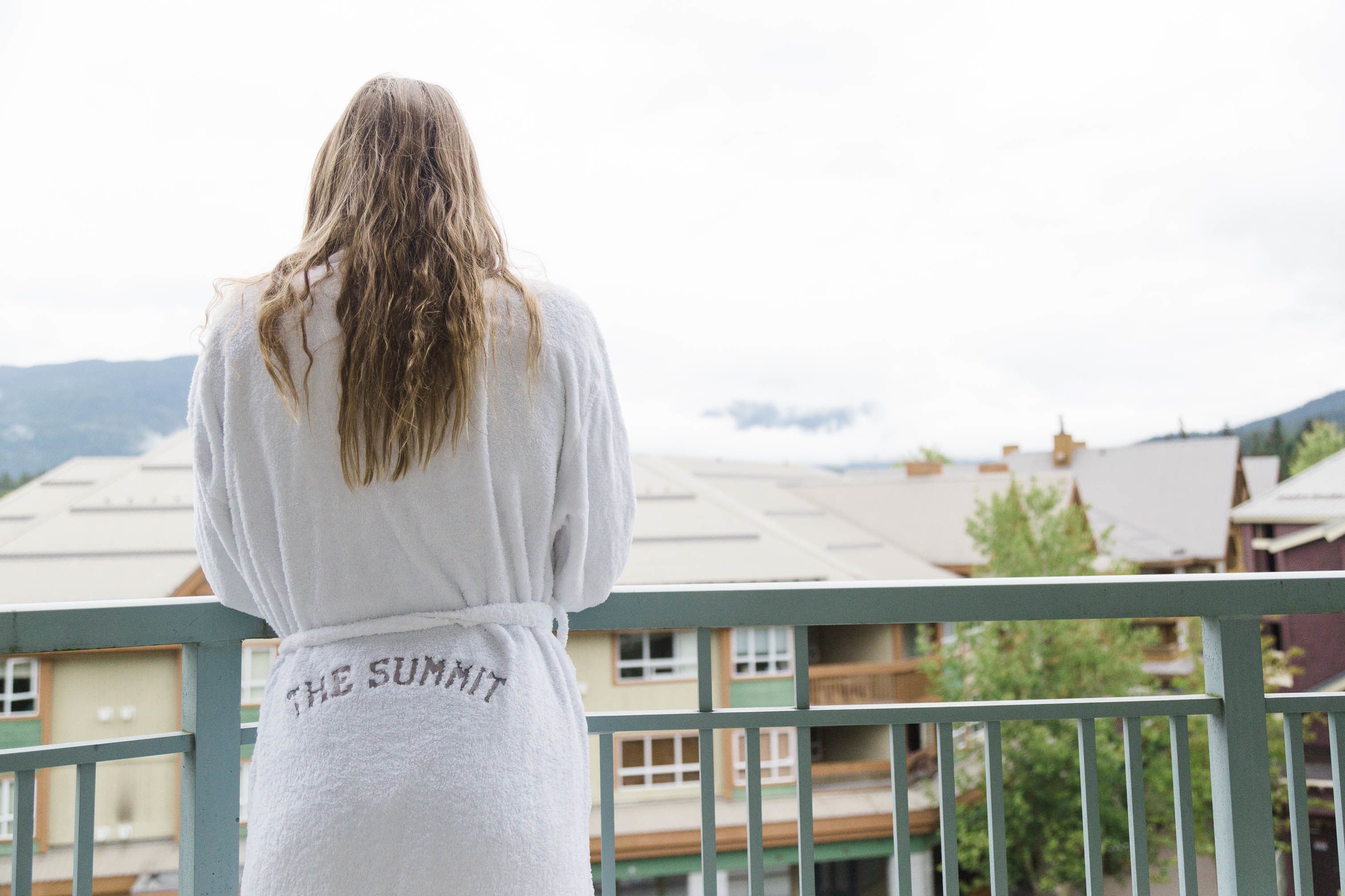 Una persona con una bata blanca bordada con la inscripción "THE SUMMIT" en la espalda, de pie en un balcón con vistas a edificios y montañas.