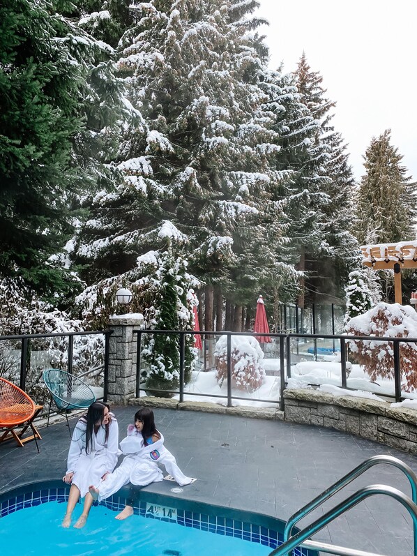 Una piscina con dos personas sentadas en el borde, rodeada de árboles cubiertos de nieve y un muro de piedra.
