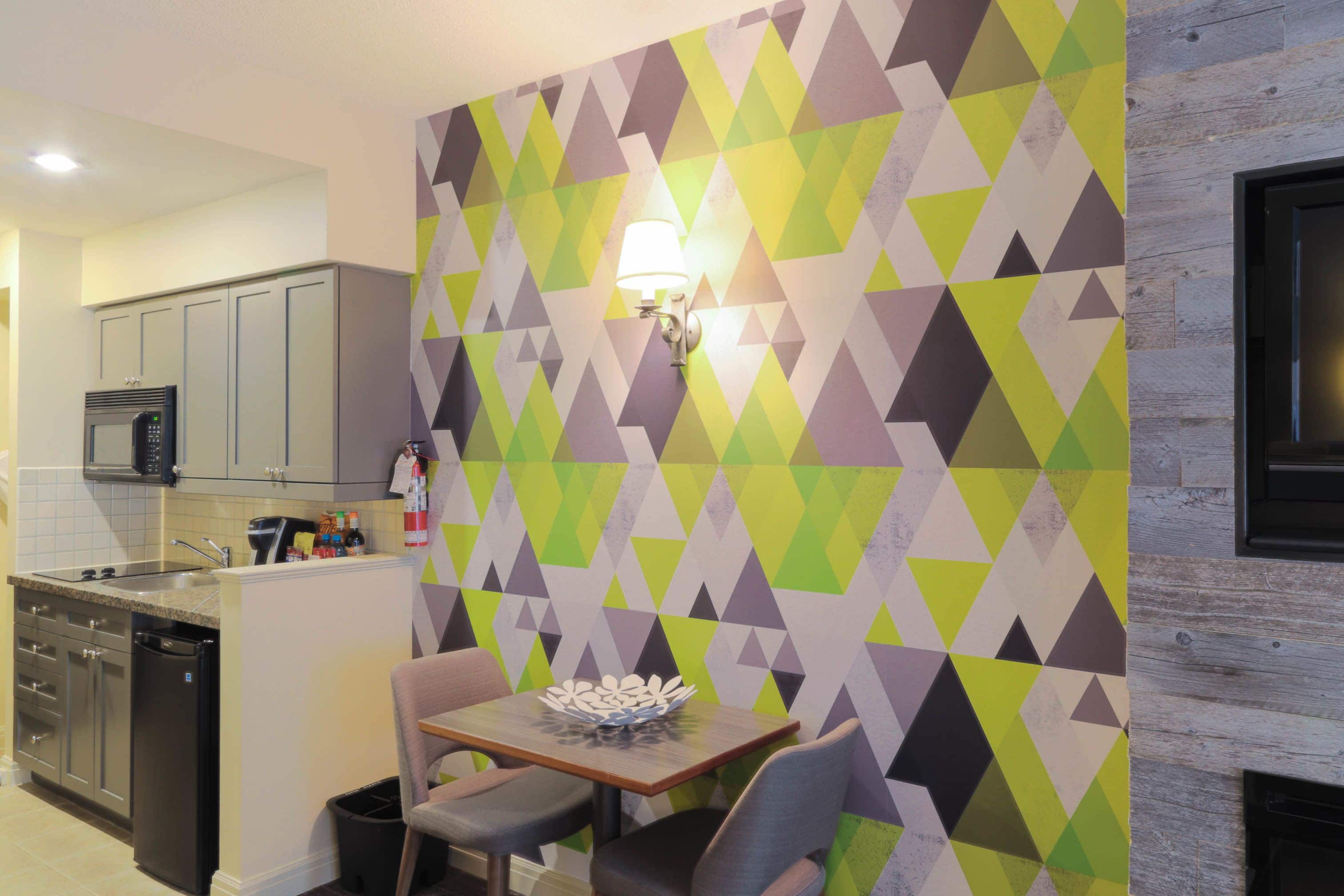 A kitchen with a dining area, featuring a geometric patterned wall, a microwave, cabinets, and a small table with chairs.