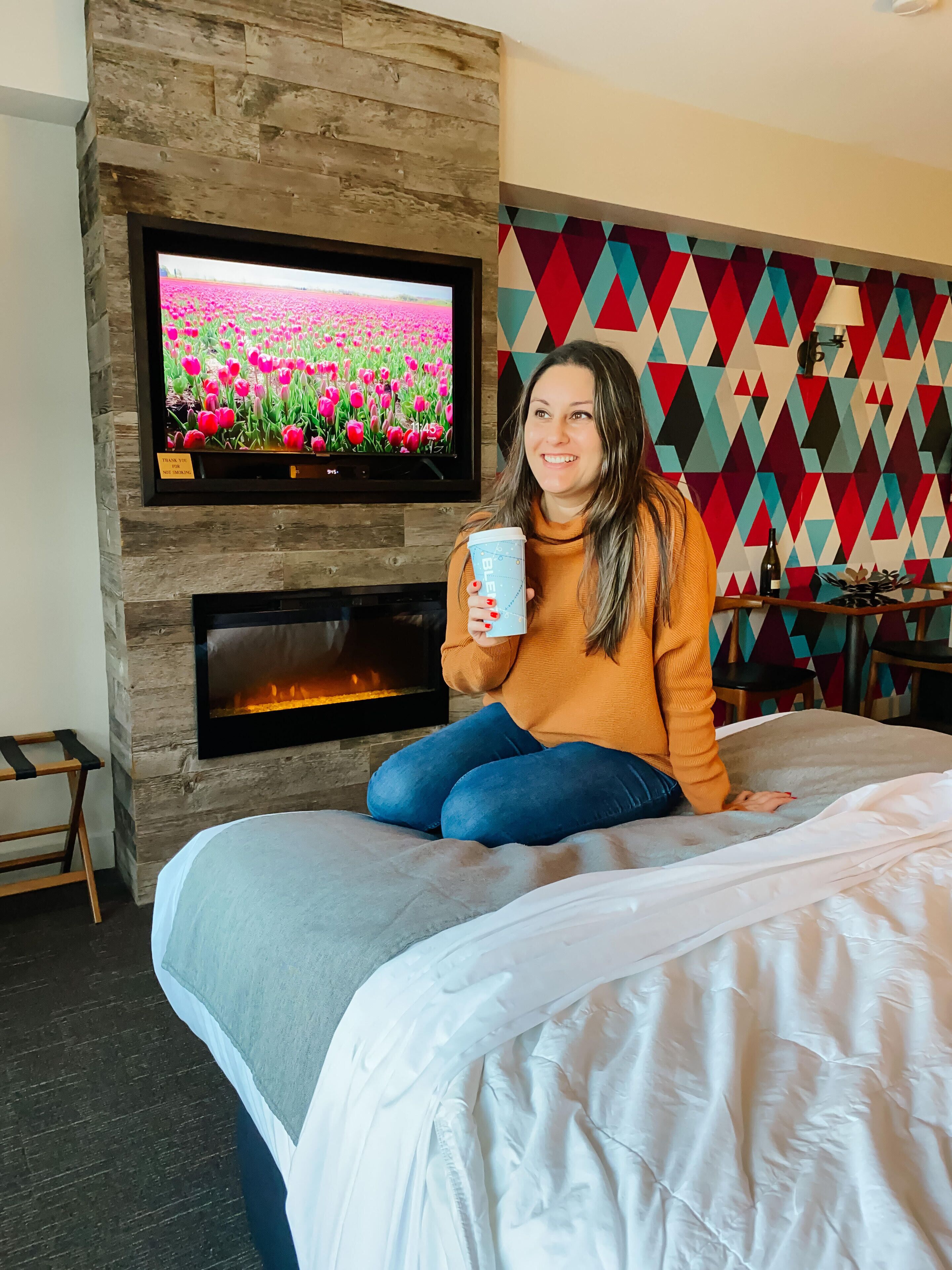 A person sitting on a bed in a room with a fireplace, a TV displaying a field of pink flowers, and a colorful geometric wall pattern.