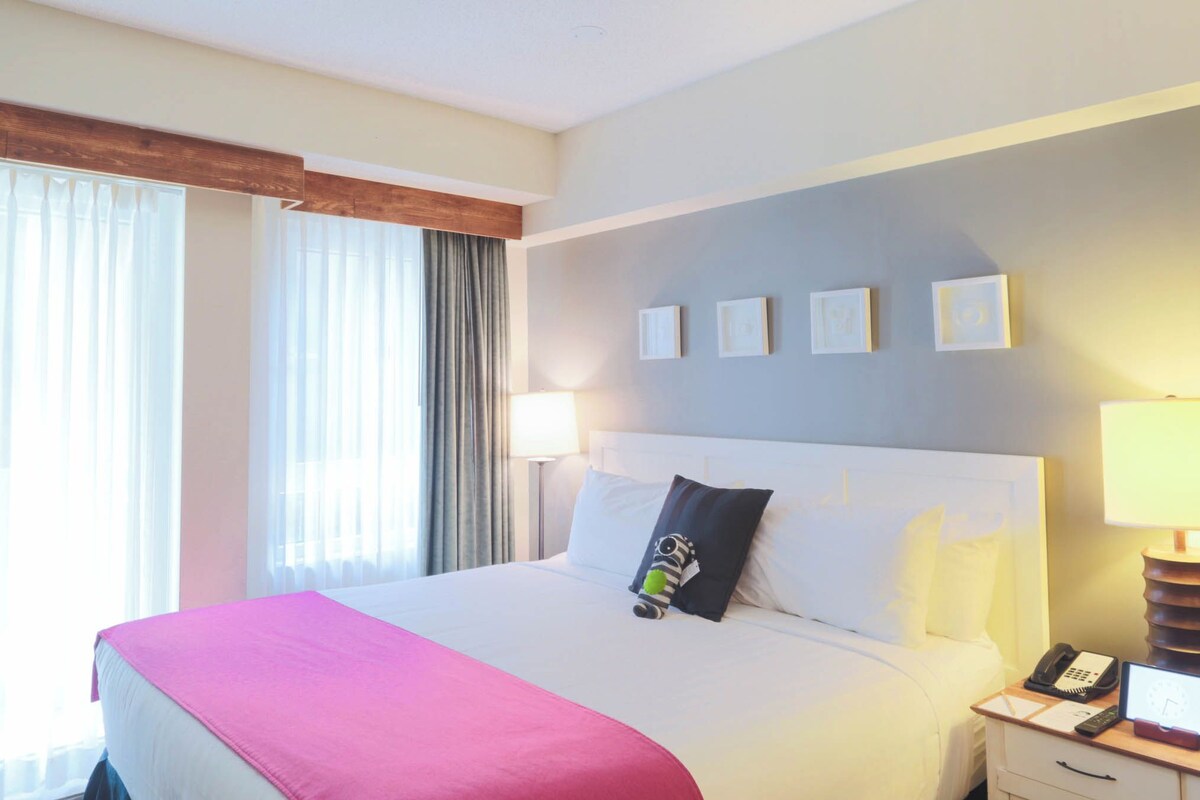 A hotel room with a bed, bedside lamps, a nightstand with a phone, and a window with curtains.