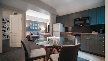A modern kitchen with dark cabinets, a microwave, and a refrigerator. There is a glass dining table with chairs in the foreground.