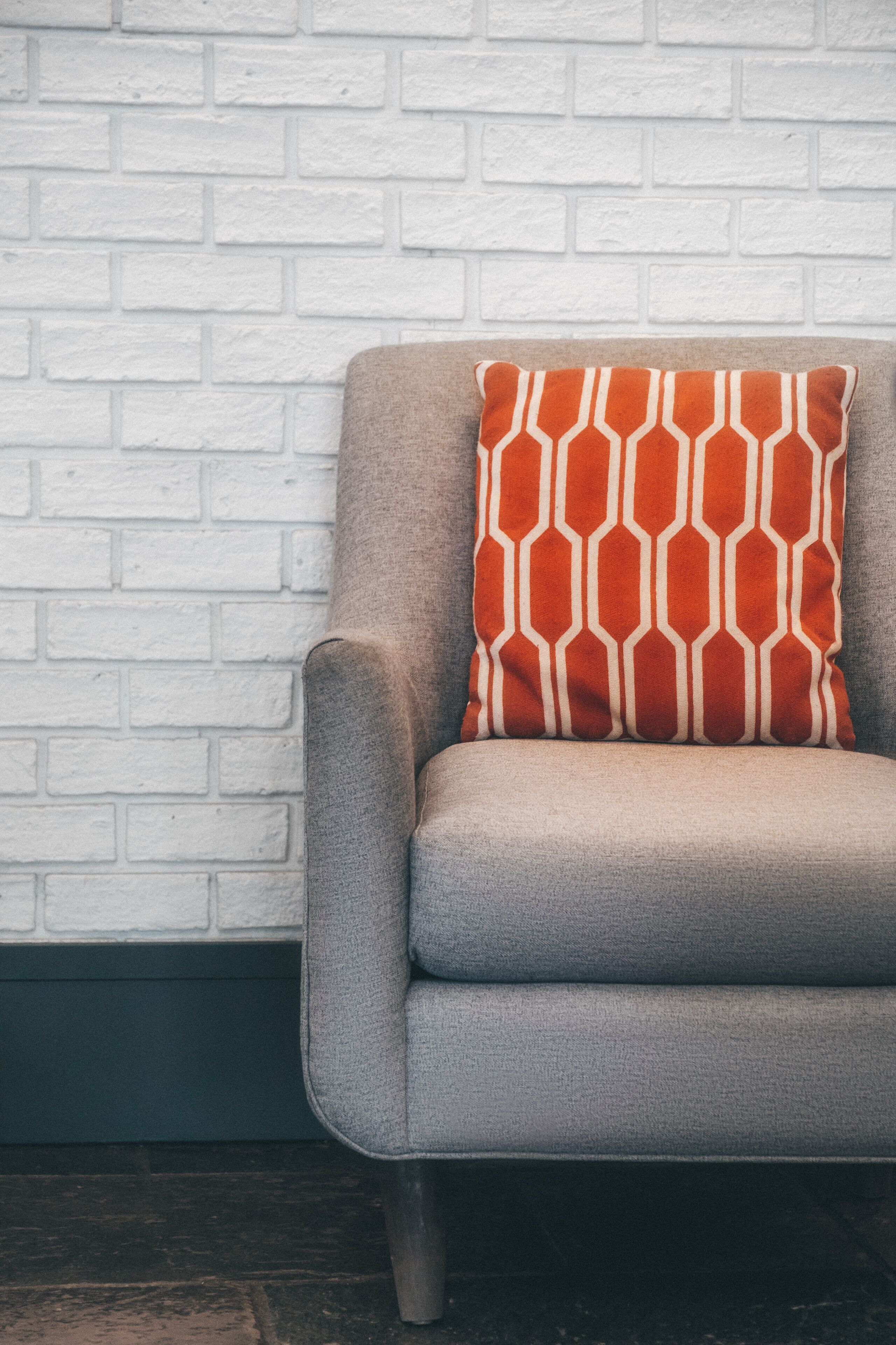A grey armchair with a patterned cushion against a white brick wall.