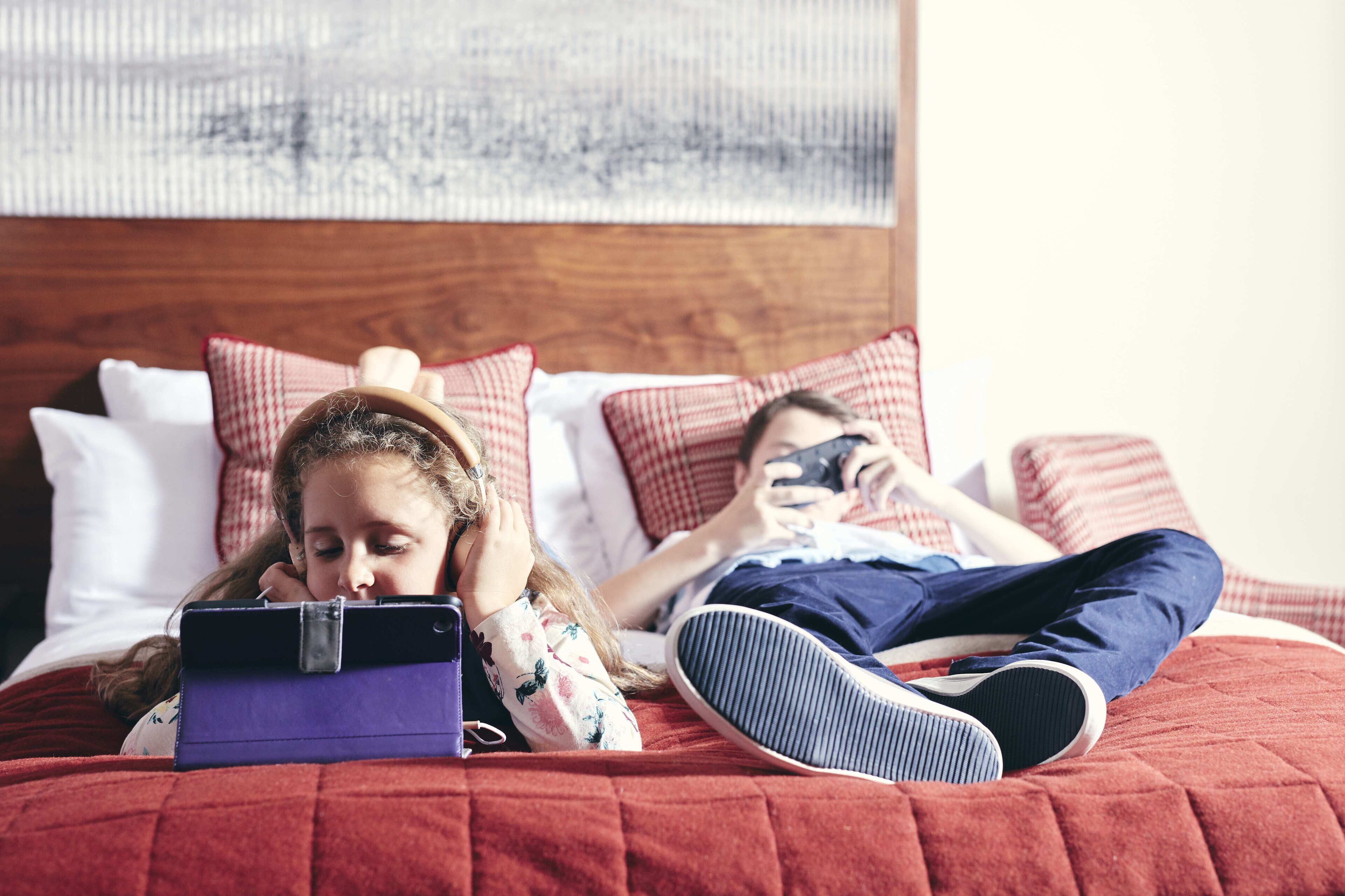 Ein Bett mit einer roten Bettdecke und einem hölzernen Kopfteil, ein violettes Tablet und ein Smartphone.