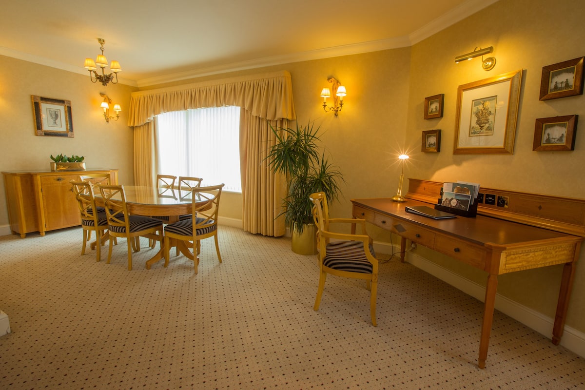 A hotel room with a dining table, chairs, a desk, a lamp, and a potted plant.