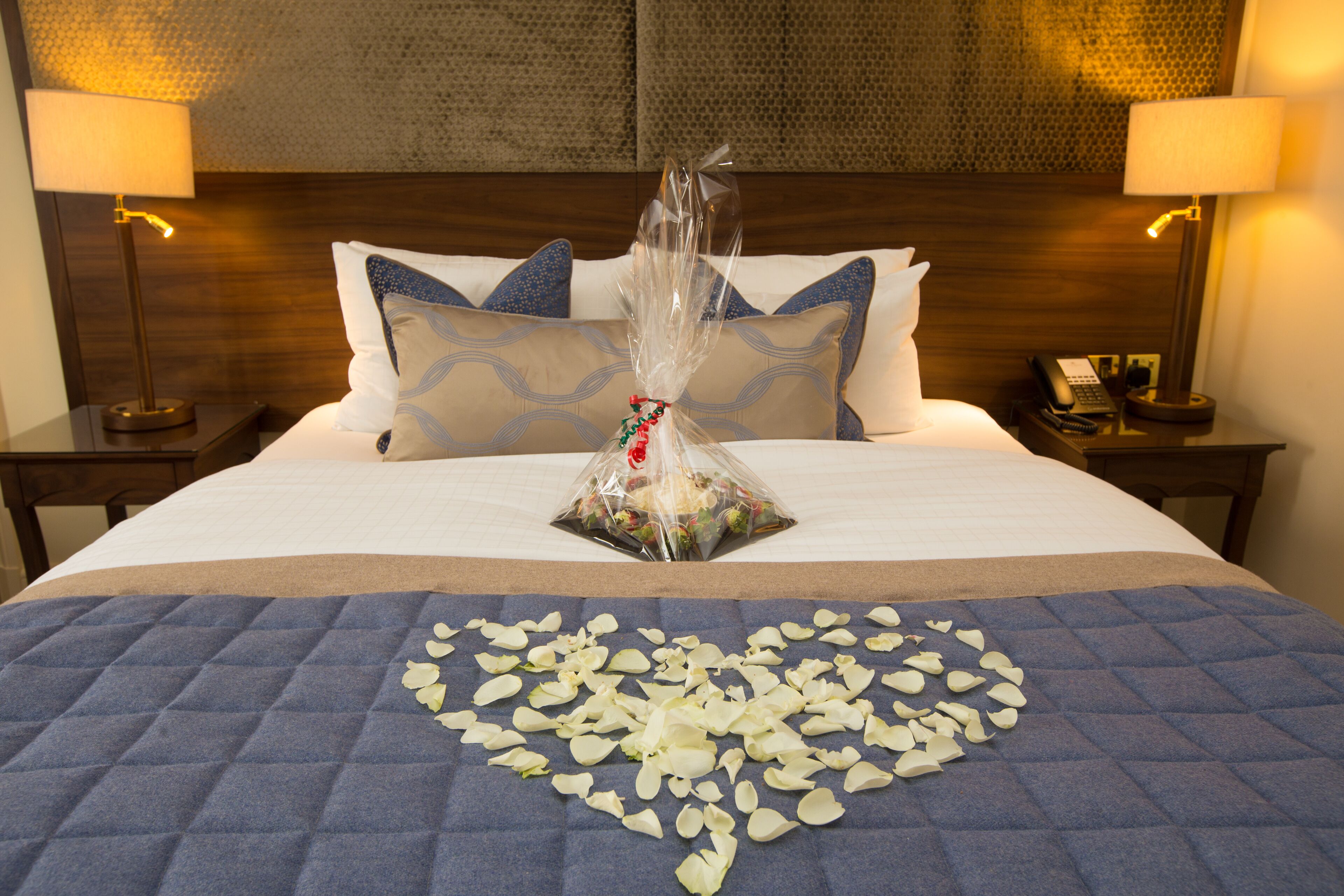 A neatly made bed with a blue quilt, white pillows, and a decorative arrangement of white flowers and a gift bag.