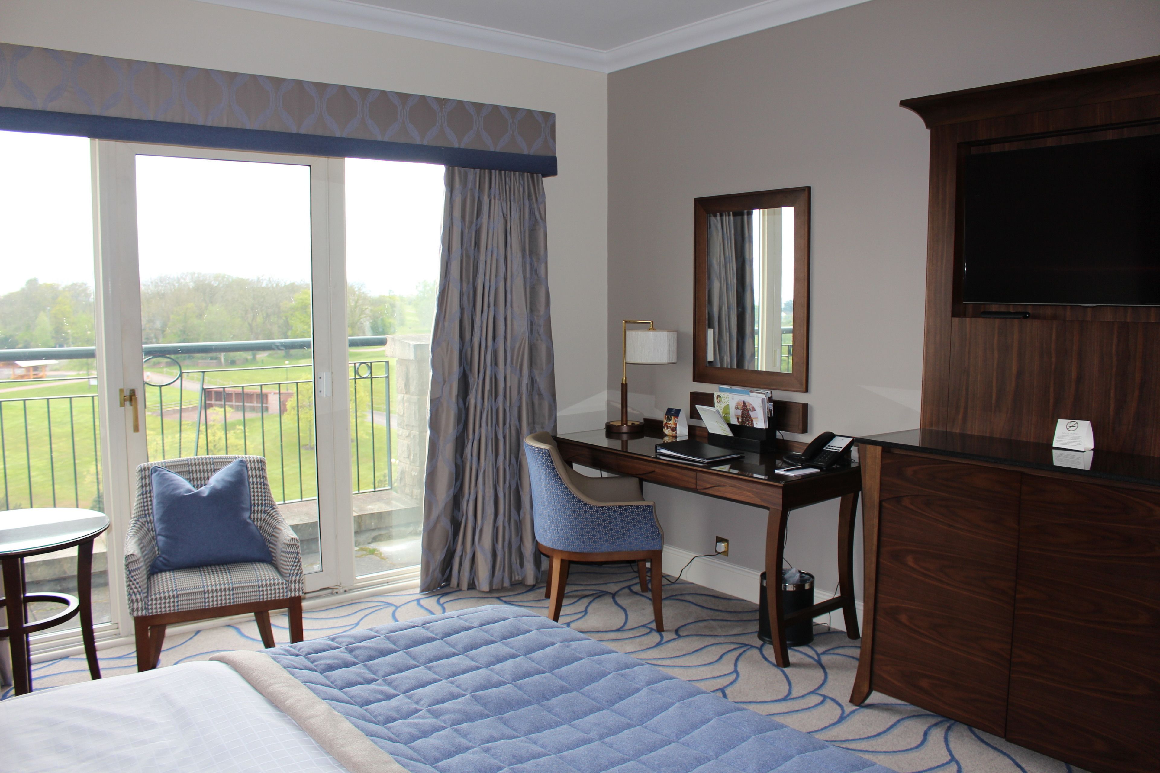 A hotel room with a bed, a desk, a chair, a TV, and a balcony with a view of greenery.