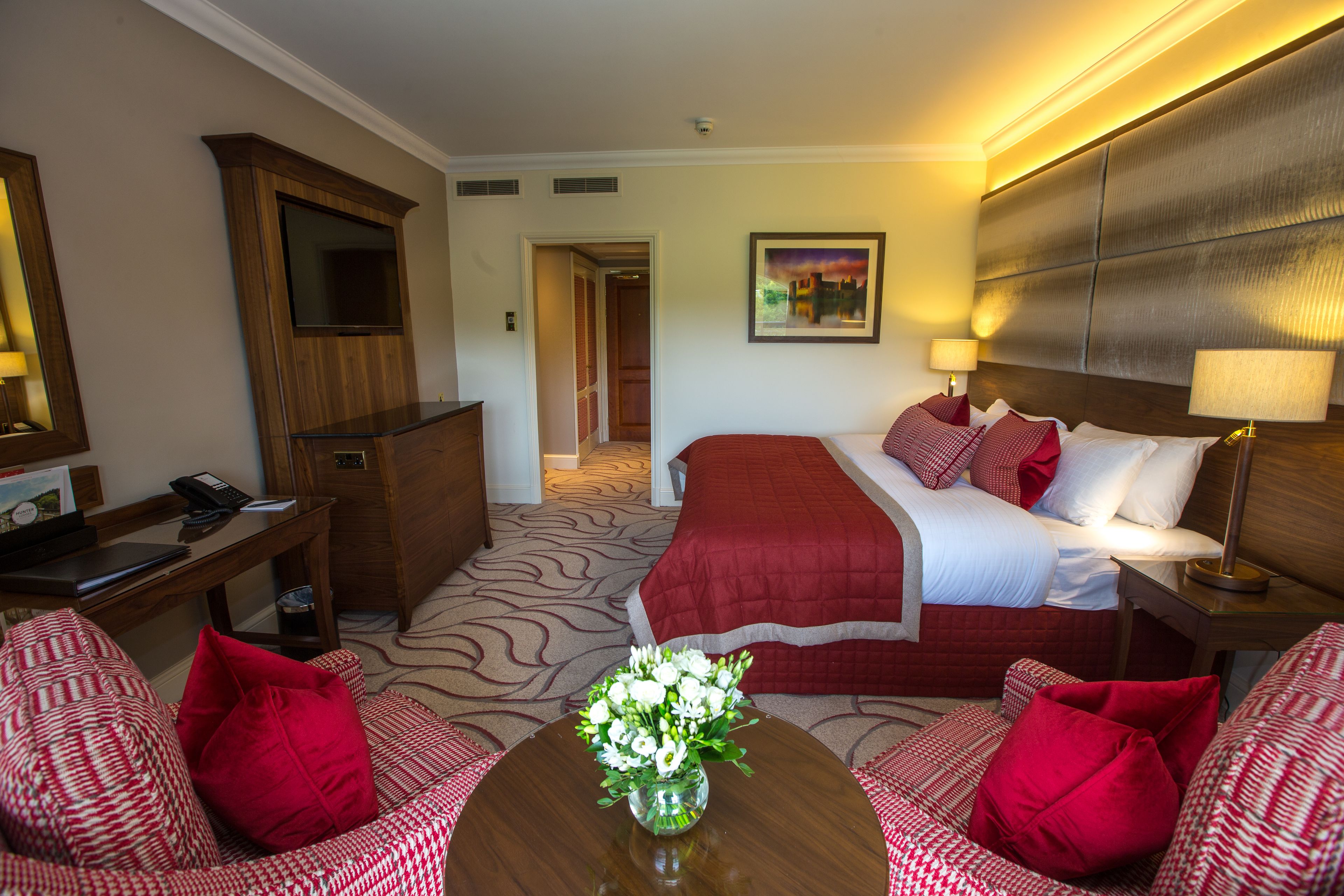Een hotelkamer met een bed, een bureau, een stoel, een bijzettafeltje met bloemen en een schilderij aan de muur.
